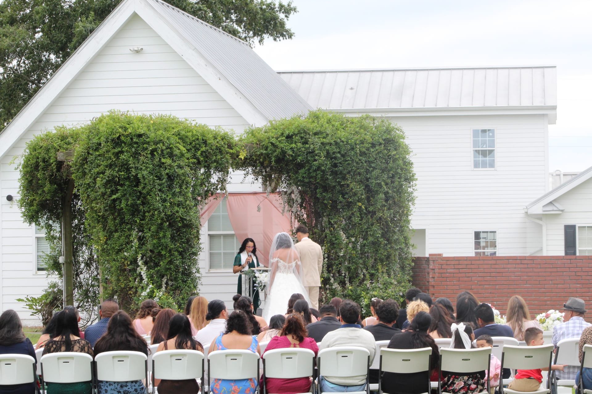 Outdoor wedding ceremony at Scarborough House with bride and groom standing beneath a lush ivy-covered arch while guests are seated in white chairs on the ceremony lawn.
