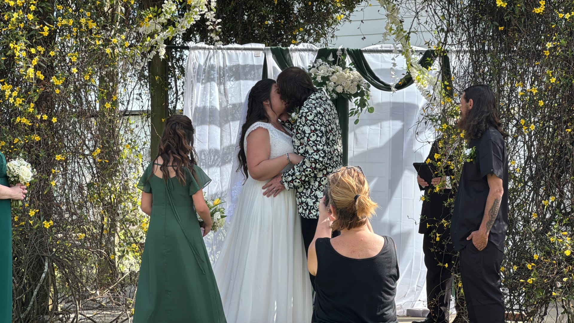 Two brides share their first kiss beneath a floral-draped ceremony arch adorned with white blooms and yellow jasmine at Scarborough House wedding venue.