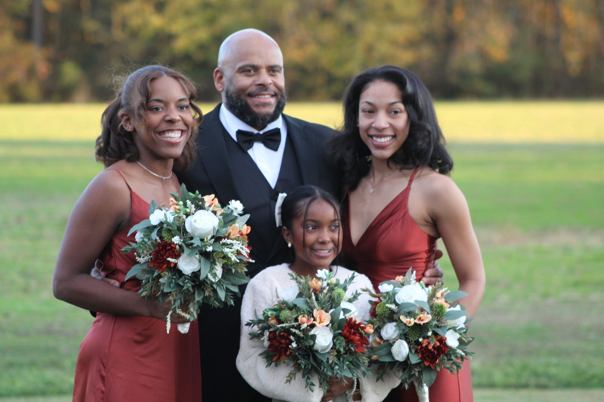 A smiling man in a tuxedo poses with two bridesmaids in rust-colored gowns and a young girl holding floral bouquets on the open lawn at Scarborough House with golden autumn trees in the background.