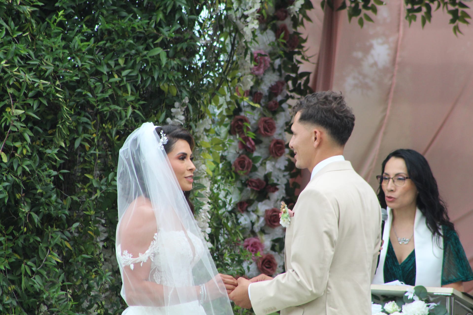 Bride in a lace gown and veil exchanges vows with groom in a cream suit while holding hands before a lush floral arch adorned with burgundy and white roses at Scarborough House.