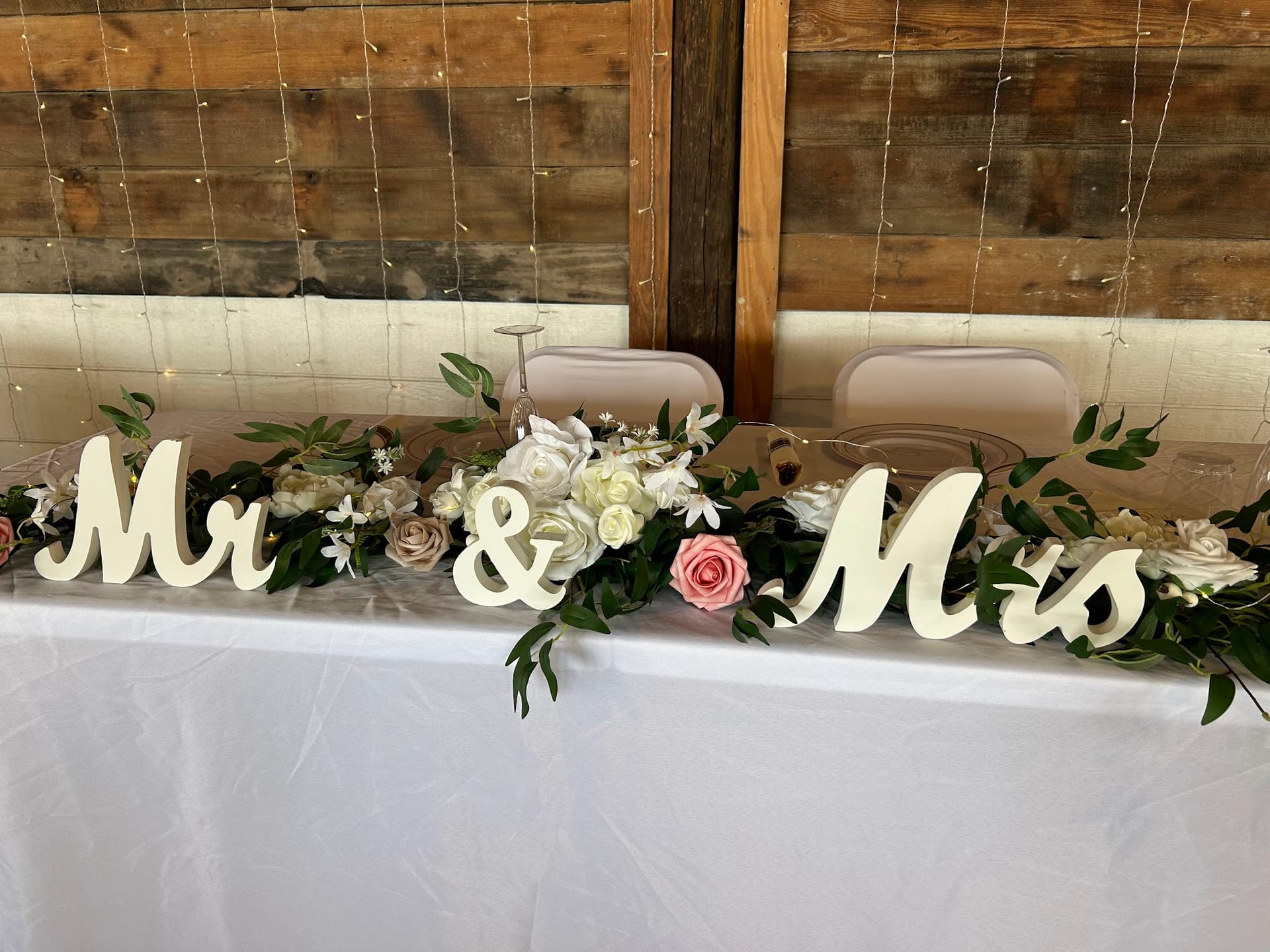 Mr and Mrs wooden signs flanking a lush floral garland of white and blush roses on a white-draped sweetheart table against a rustic wood-plank wall with string lights at Scarborough House.