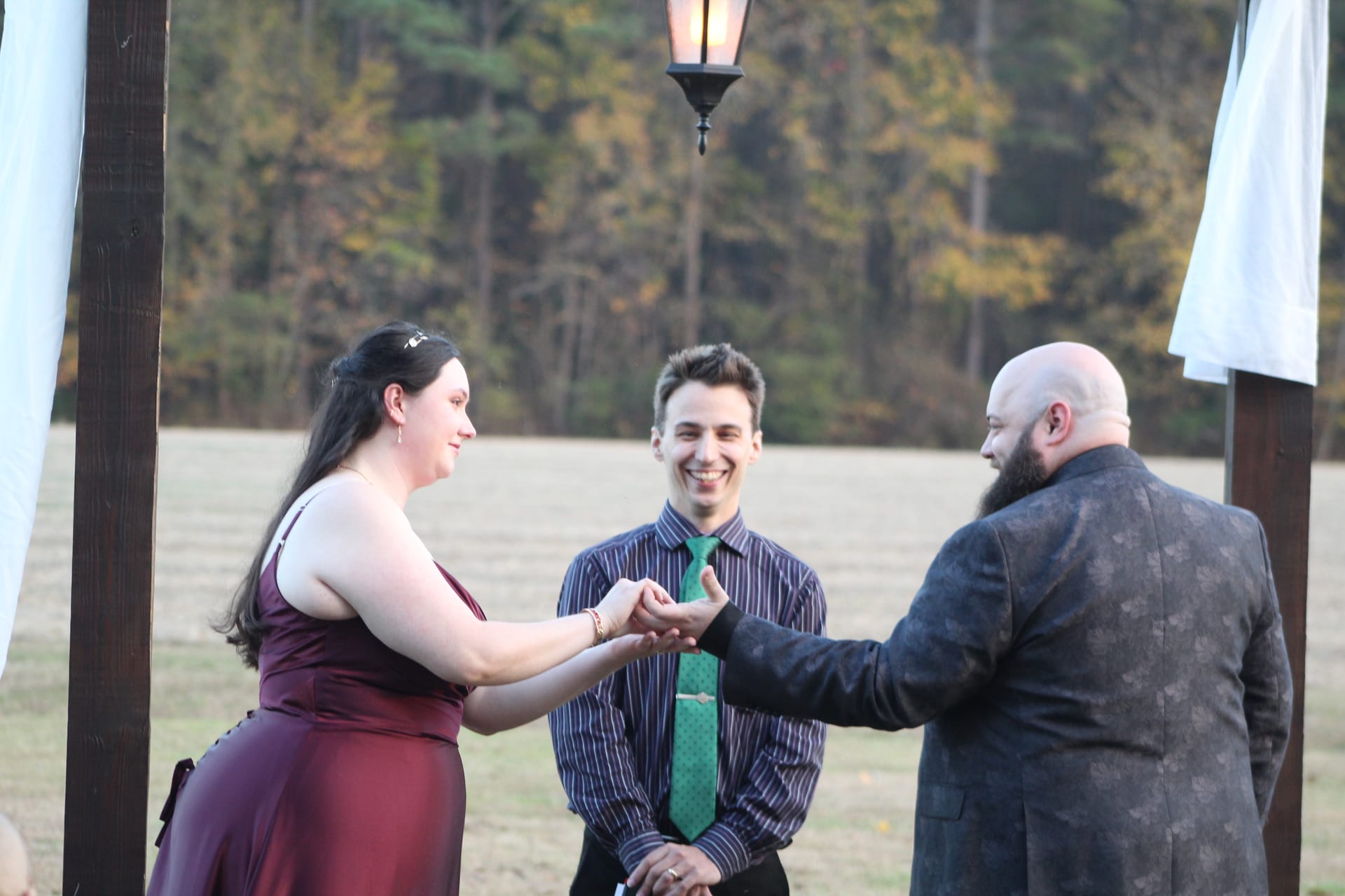 Bride in burgundy gown exchanges rings with bearded groom beneath a wooden arch with hanging lantern as a smiling officiant in a green tie looks on during an outdoor autumn wedding ceremony at Scarborough House.