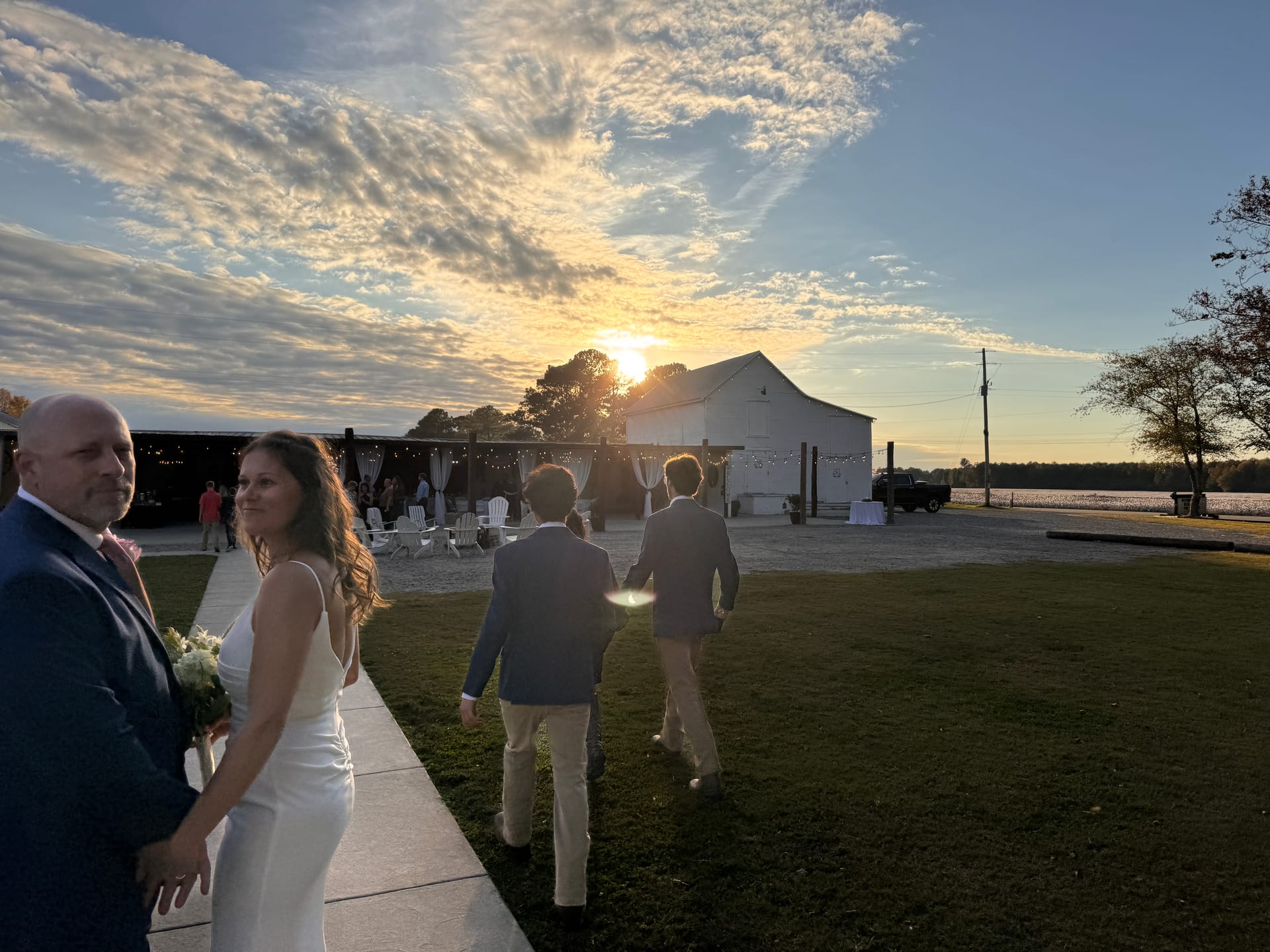 Bride in white gown glances back while walking with guests toward a white barn wedding venue bathed in a stunning golden sunset over the water.