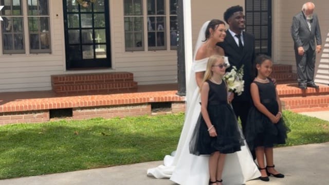 Bride in off-shoulder white gown and groom pose with two flower girls in black dresses on the lawn in front of the historic 1821 Scarborough House manor.