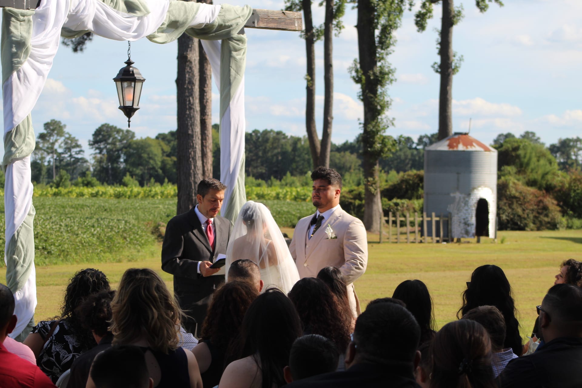 Outdoor wedding ceremony at Scarborough House with a decorated wooden arch draped in white fabric and sage green accents, a groom in a tan suit facing the veiled bride as guests watch from their seats with lush farmland fields and trees in the background.