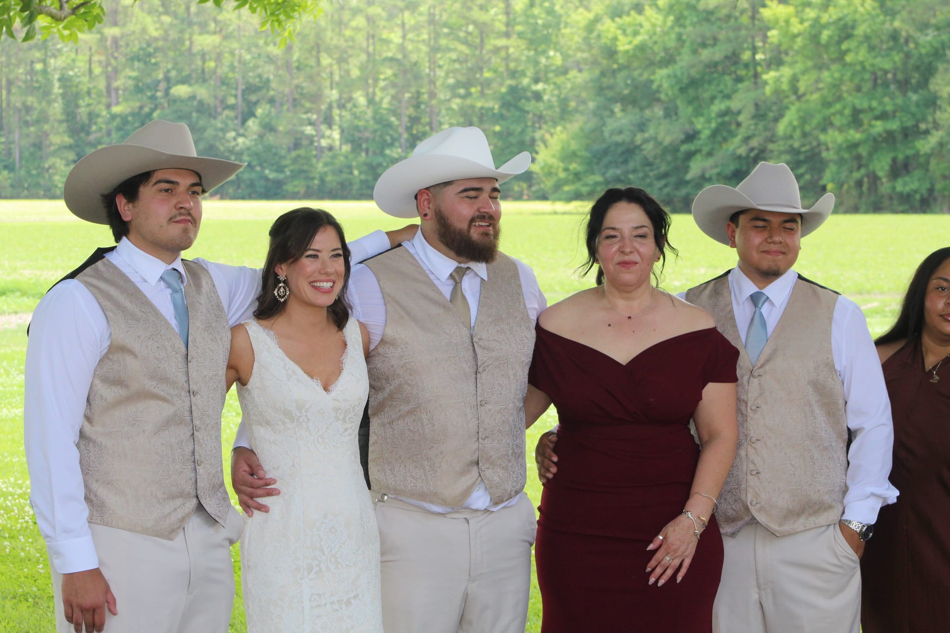 Bride in white gown and groom in tan vest and cowboy hat pose with family members in matching western attire on a green lawn surrounded by trees at Scarborough House.