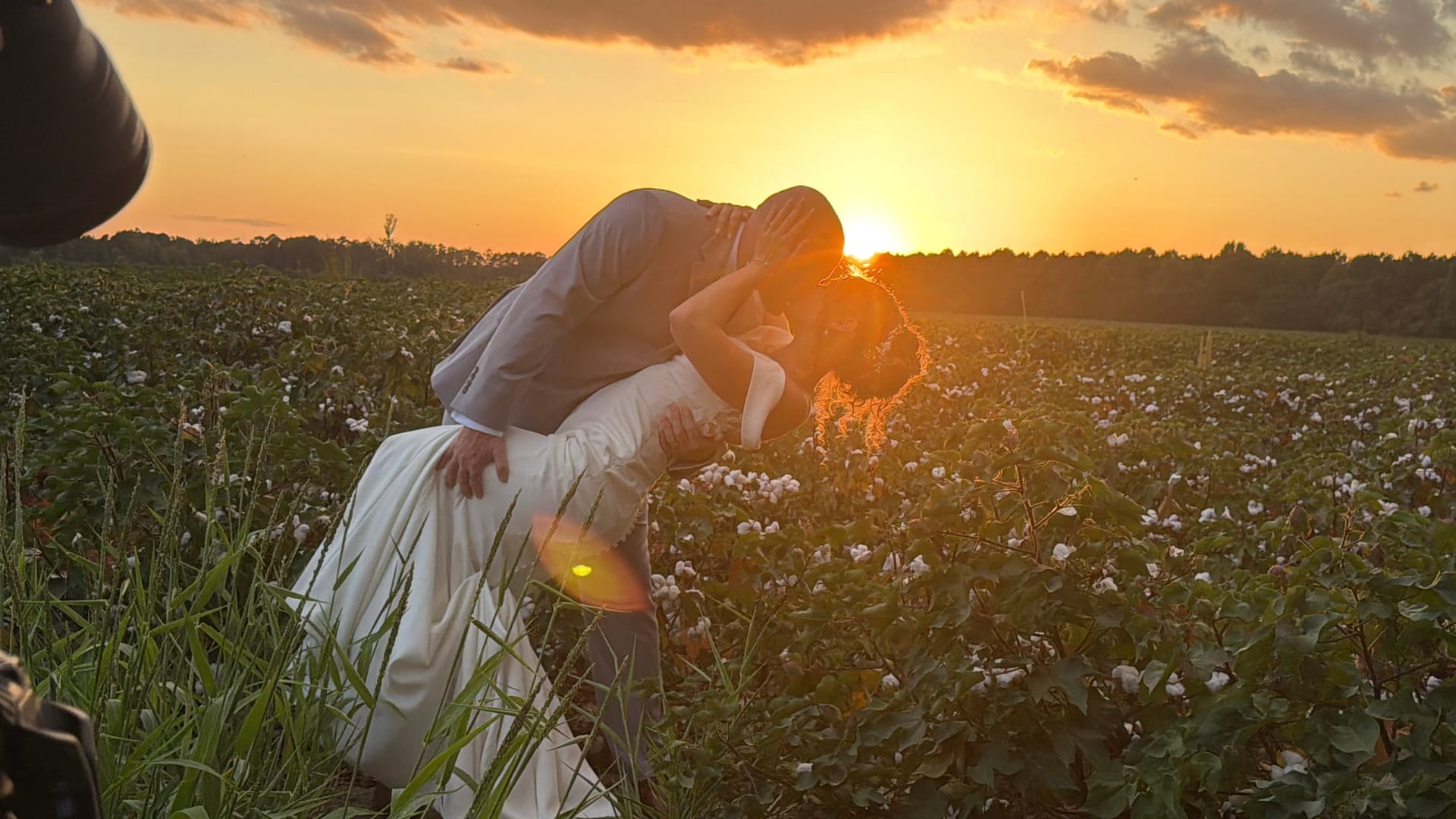 Bride and groom share a romantic dip kiss in a cotton field at golden hour with a glowing sunset horizon behind them.