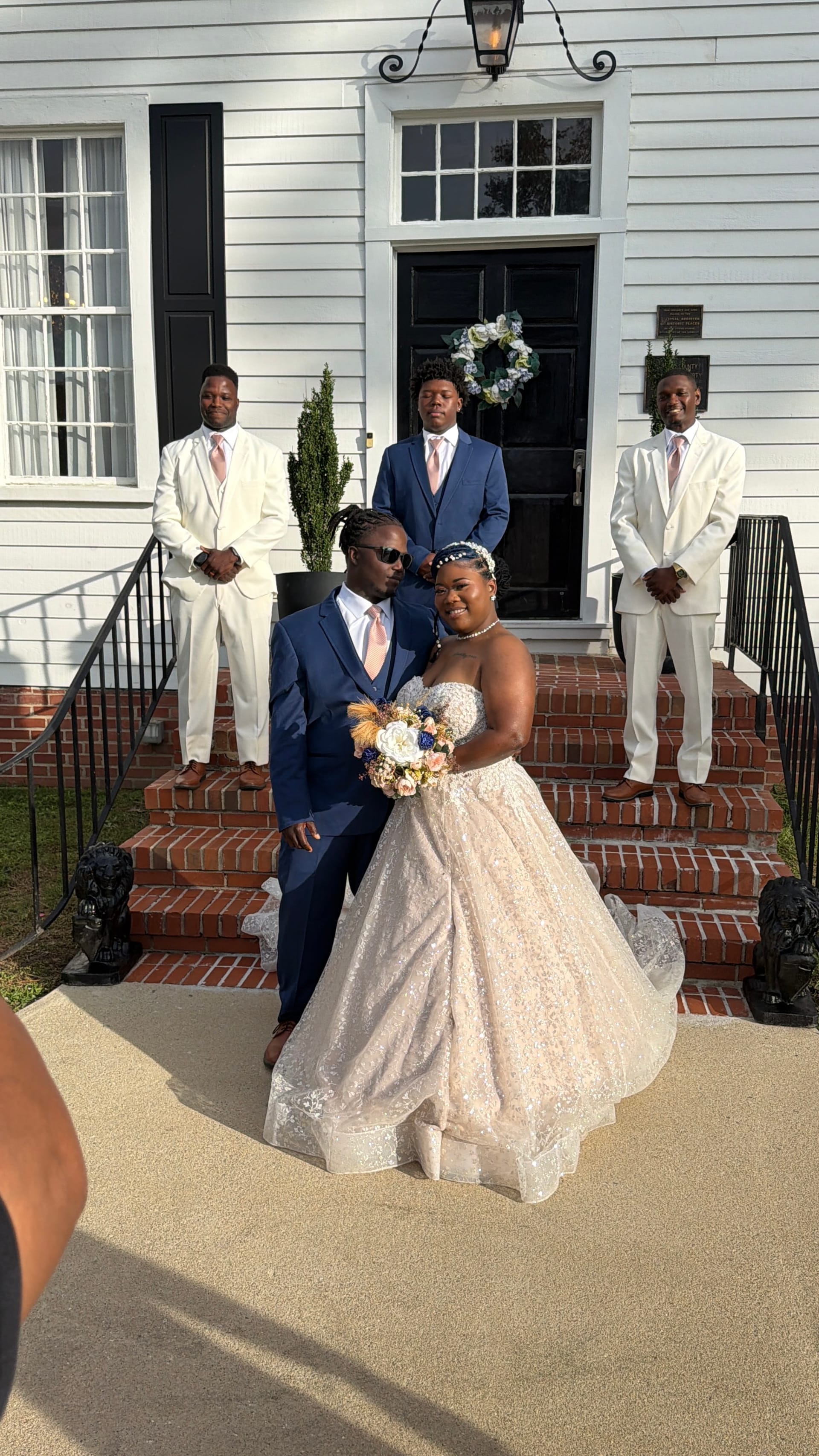 Bride in a blush ballgown and groom in a navy suit pose together on the brick front steps of the 1821 Scarborough House manor while three groomsmen in white and navy suits stand behind them.