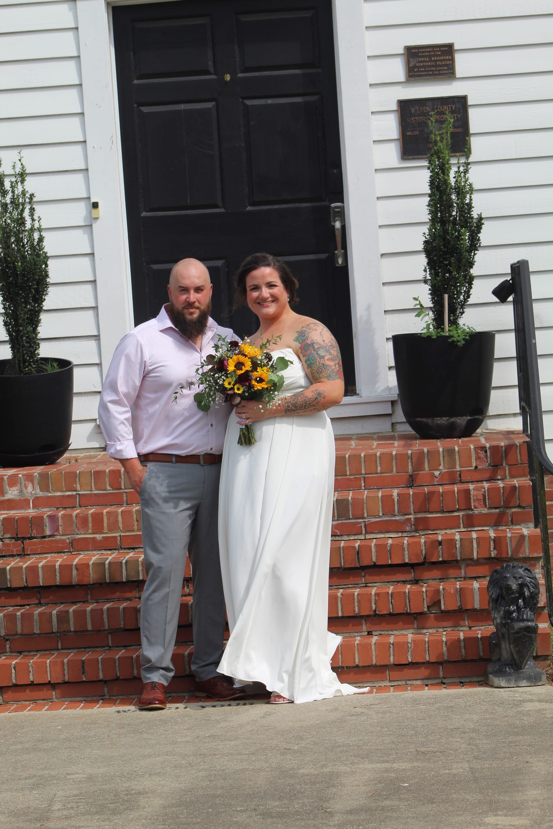 Smiling bride in a white gown holding a sunflower bouquet stands with her bearded groom in a lavender shirt on the brick front steps of the historic 1821 Scarborough House estate.