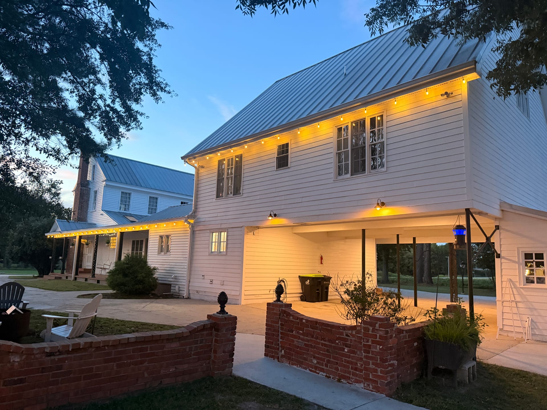 Scarborough House historic white two-story manor glowing with warm string lights at dusk, framed by a brick garden wall and lush oak trees.