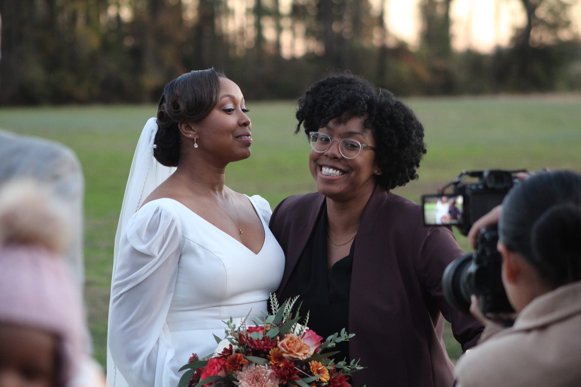 A smiling bride in a white long-sleeve gown holding a colorful autumn bouquet shares a warm moment with a joyful guest in a burgundy blazer on the ceremony lawn at Scarborough House.