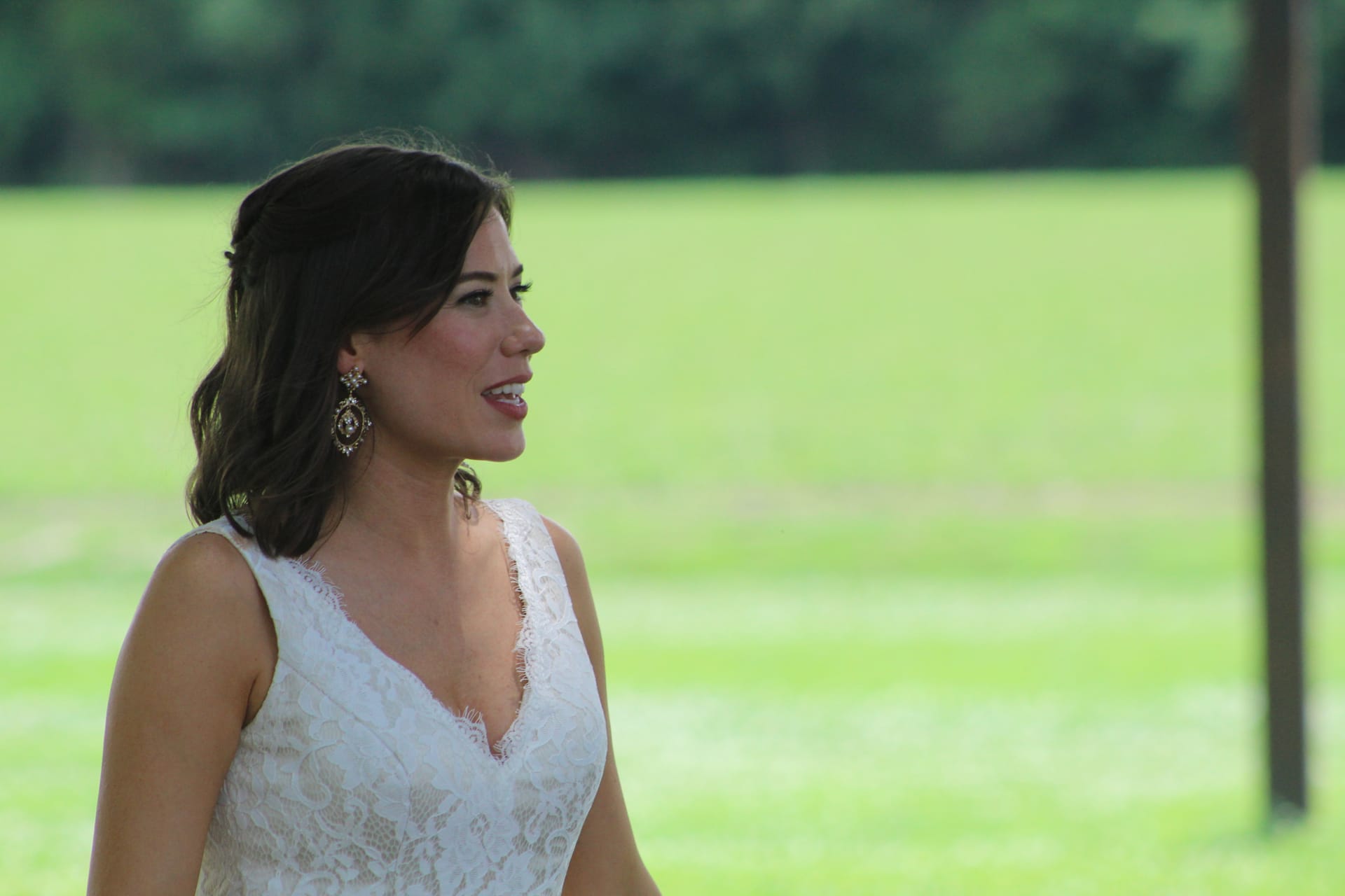Bride in a white lace gown smiling graciously in profile against a lush green lawn and tree backdrop at Scarborough House estate.