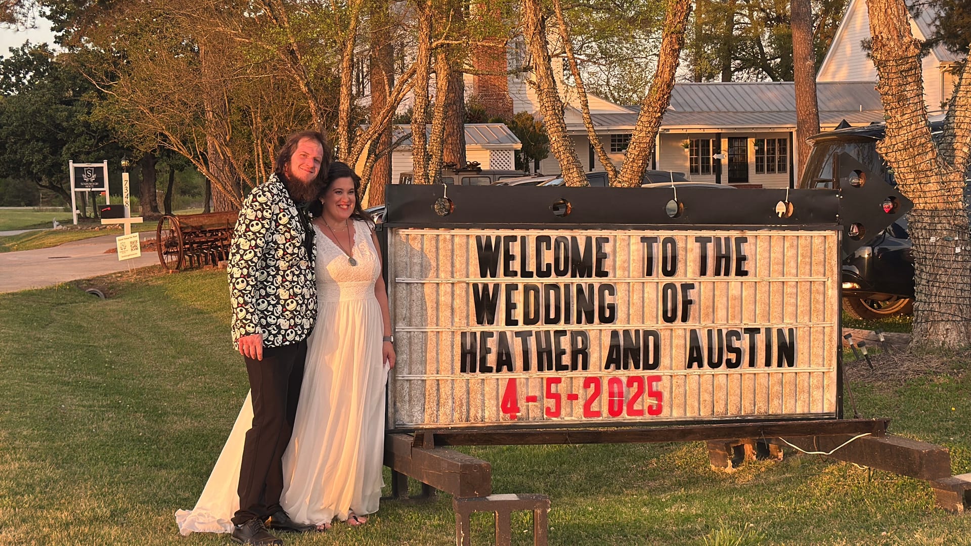Bride in a flowing white gown and groom in a floral blazer smile together beside a lit marquee sign welcoming guests to the wedding of Heather and Austin on April 5 2025 at a rustic estate venue during golden hour.