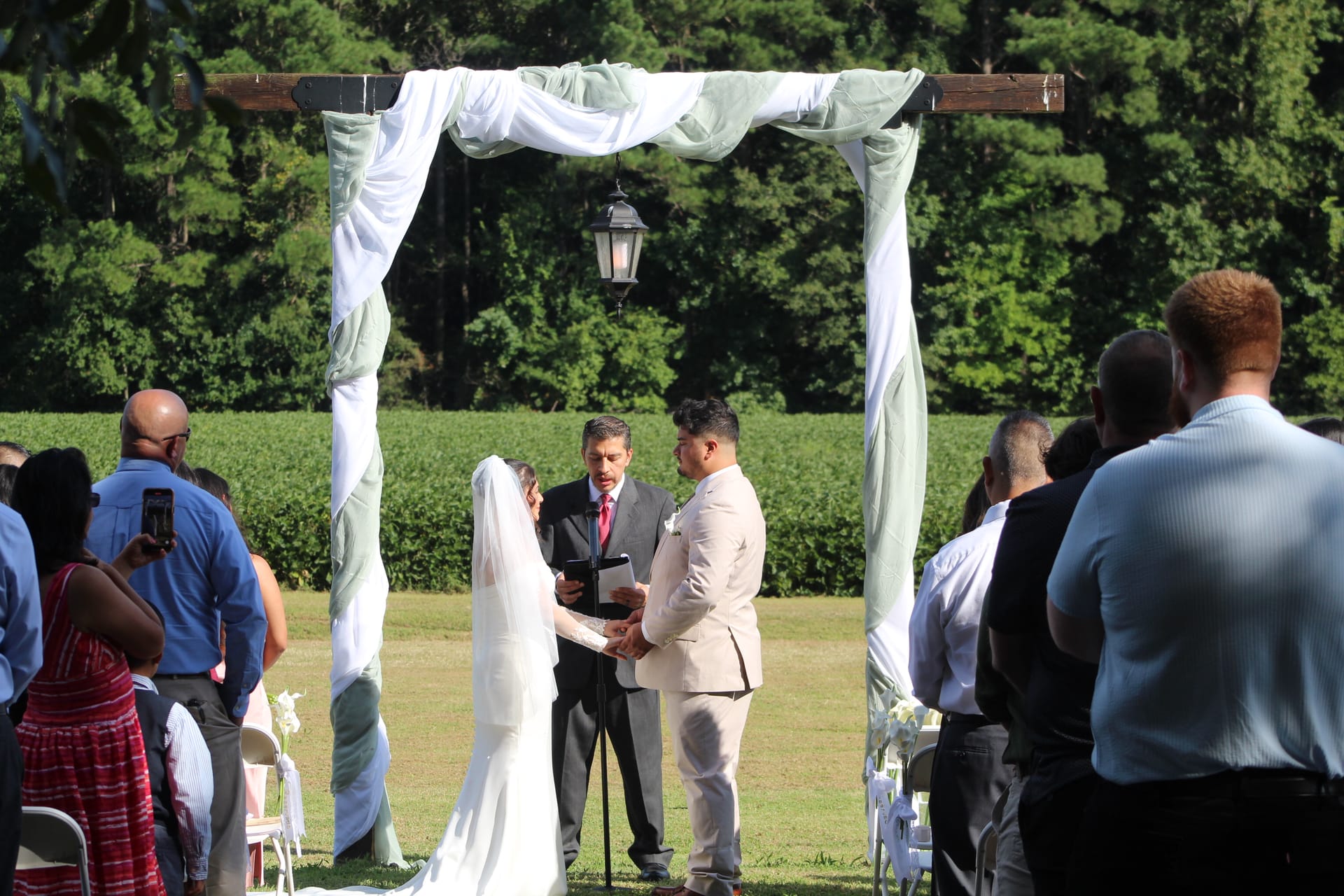 A bride and groom hold hands beneath a rustic wooden arch draped in white and sage fabric with a hanging lantern as their officiant reads vows on the ceremony lawn at Scarborough House, surrounded by lush green farmland fields and gathered guests.
