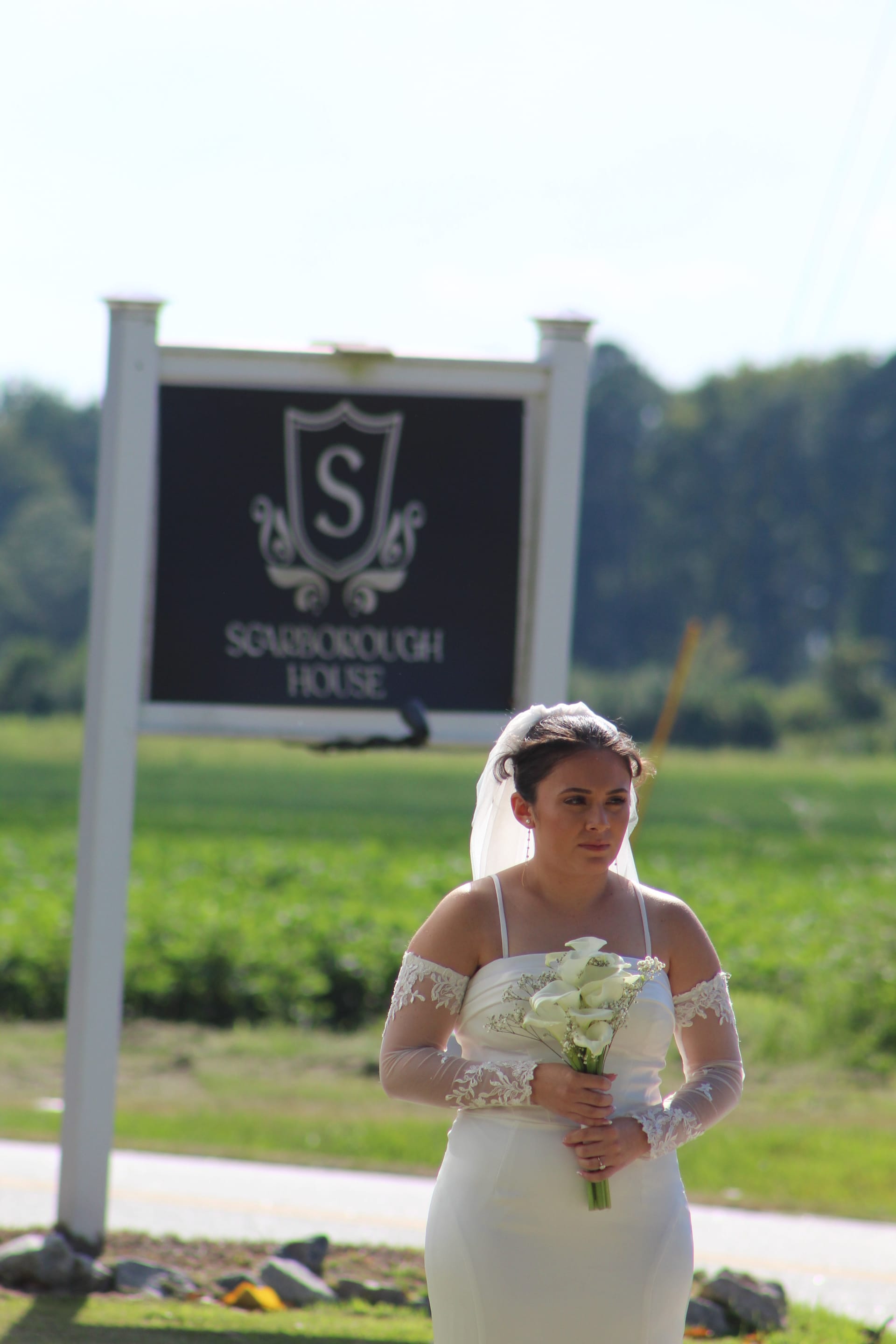 Bride in a fitted white gown and lace sleeves holding a white calla lily bouquet standing in front of the Scarborough House estate sign on a sunny day.