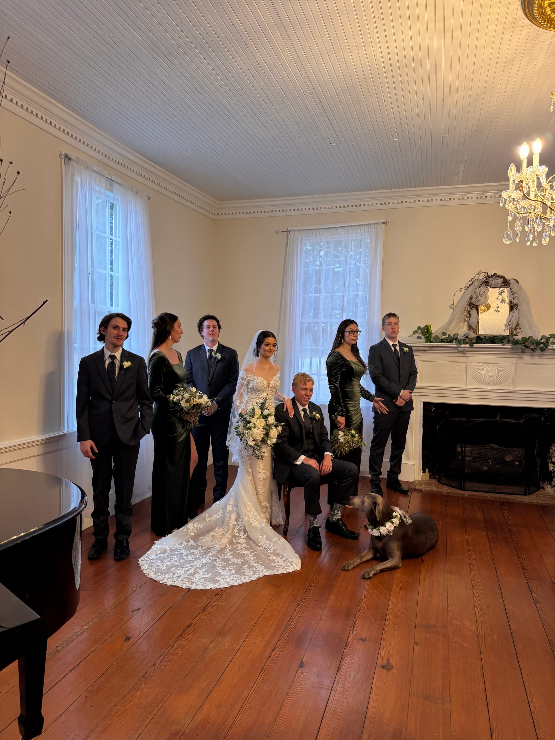 Bride in lace gown and groom seated with their wedding party dressed in black posing beside a fireplace in the historic 1821 grand hall of Scarborough House, with their dog wearing a floral collar lying at their feet.