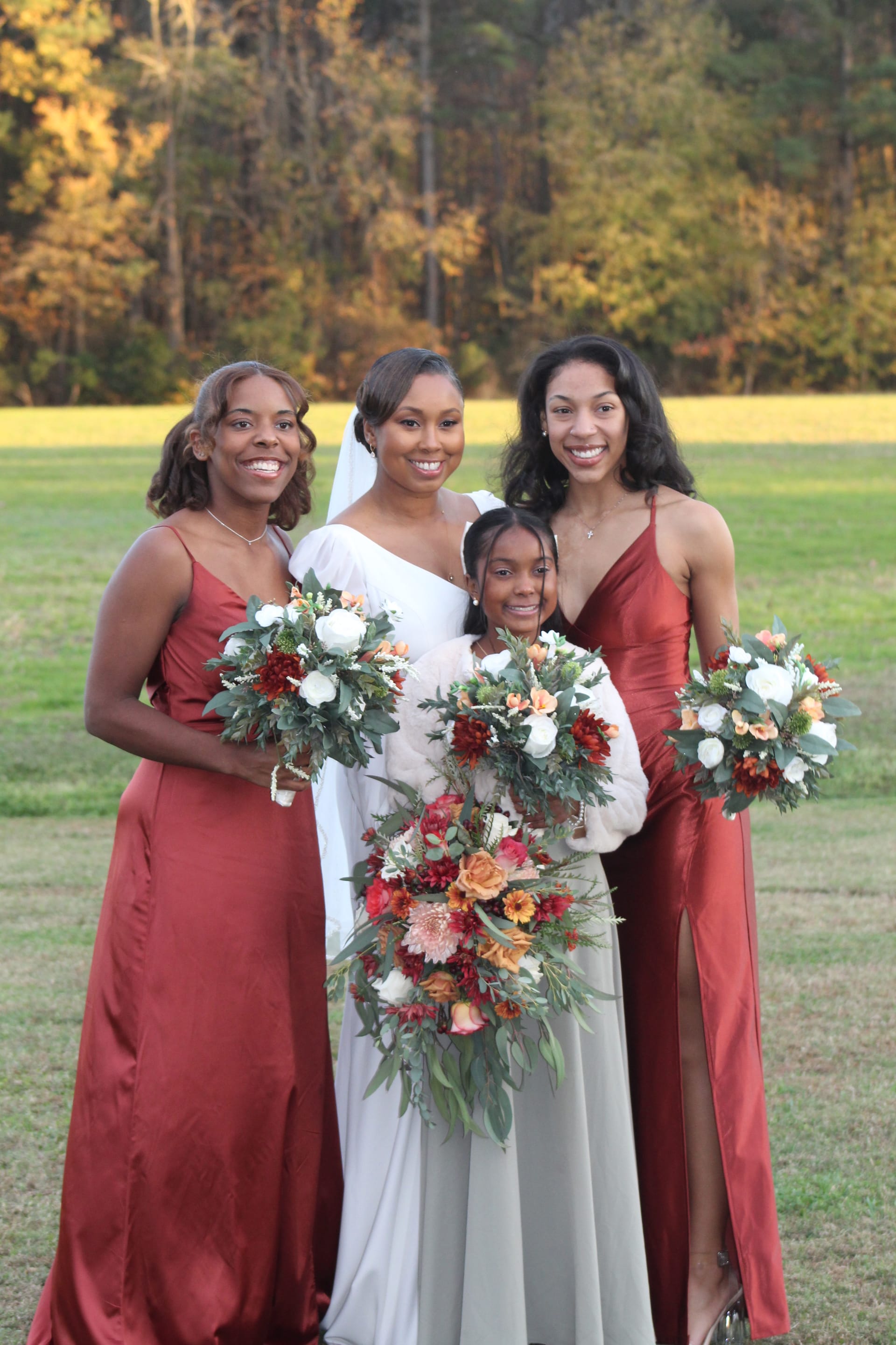 Bride in white gown with veil and two bridesmaids in rust-red satin dresses holding autumn floral bouquets on the ceremony lawn at Scarborough House with golden fall foliage in the background.
