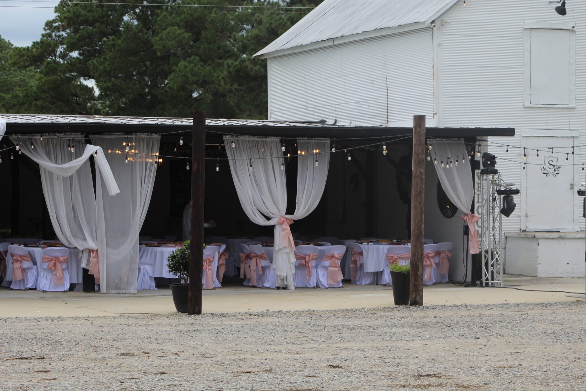 Outdoor magnolia pavilion at Scarborough House dressed for a wedding reception with white chair covers, blush pink sashes, sheer draped curtains, and warm string lights against a rustic white barn backdrop.