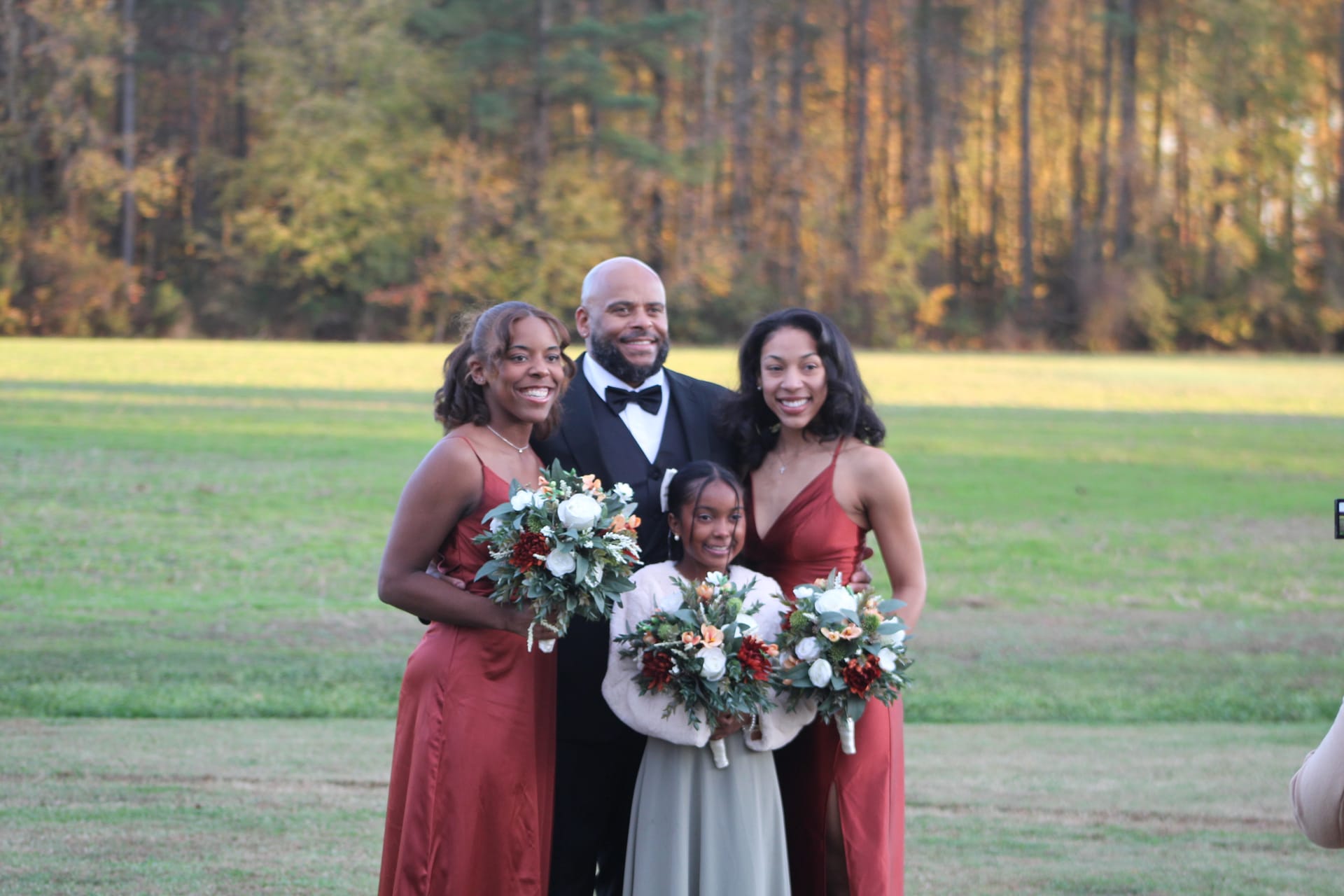 A man in a tuxedo poses with two bridesmaids in rust-colored satin gowns holding bouquets of white and burgundy flowers and a young girl in a white dress against an autumn field backdrop at Scarborough House.