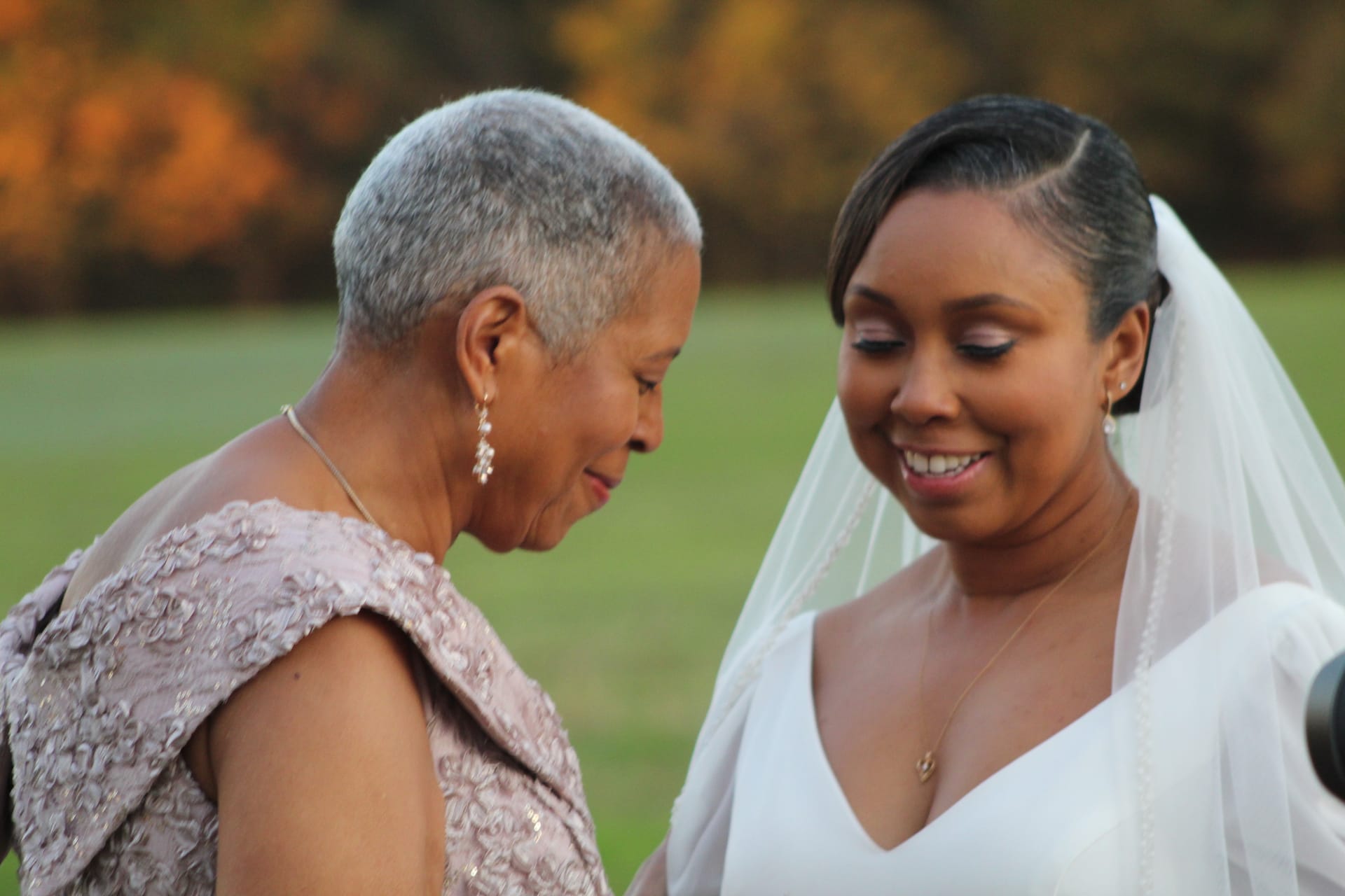 A smiling bride in a white gown and veil shares a warm candid moment with an older woman in a blush lace dress against a golden autumn backdrop at Scarborough House.