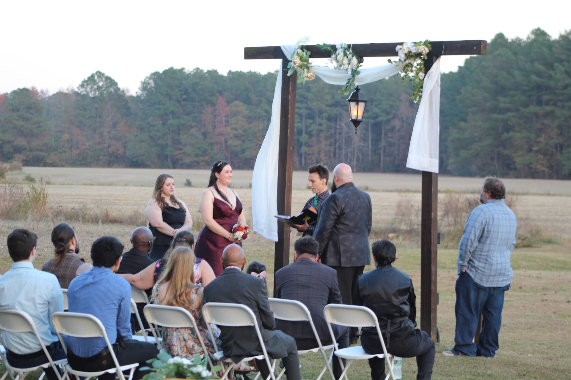 A small outdoor wedding ceremony underway beneath a rustic wooden arch draped in white fabric and flowers, with guests seated on folding chairs facing the couple and officiant against an open farmland backdrop at dusk.