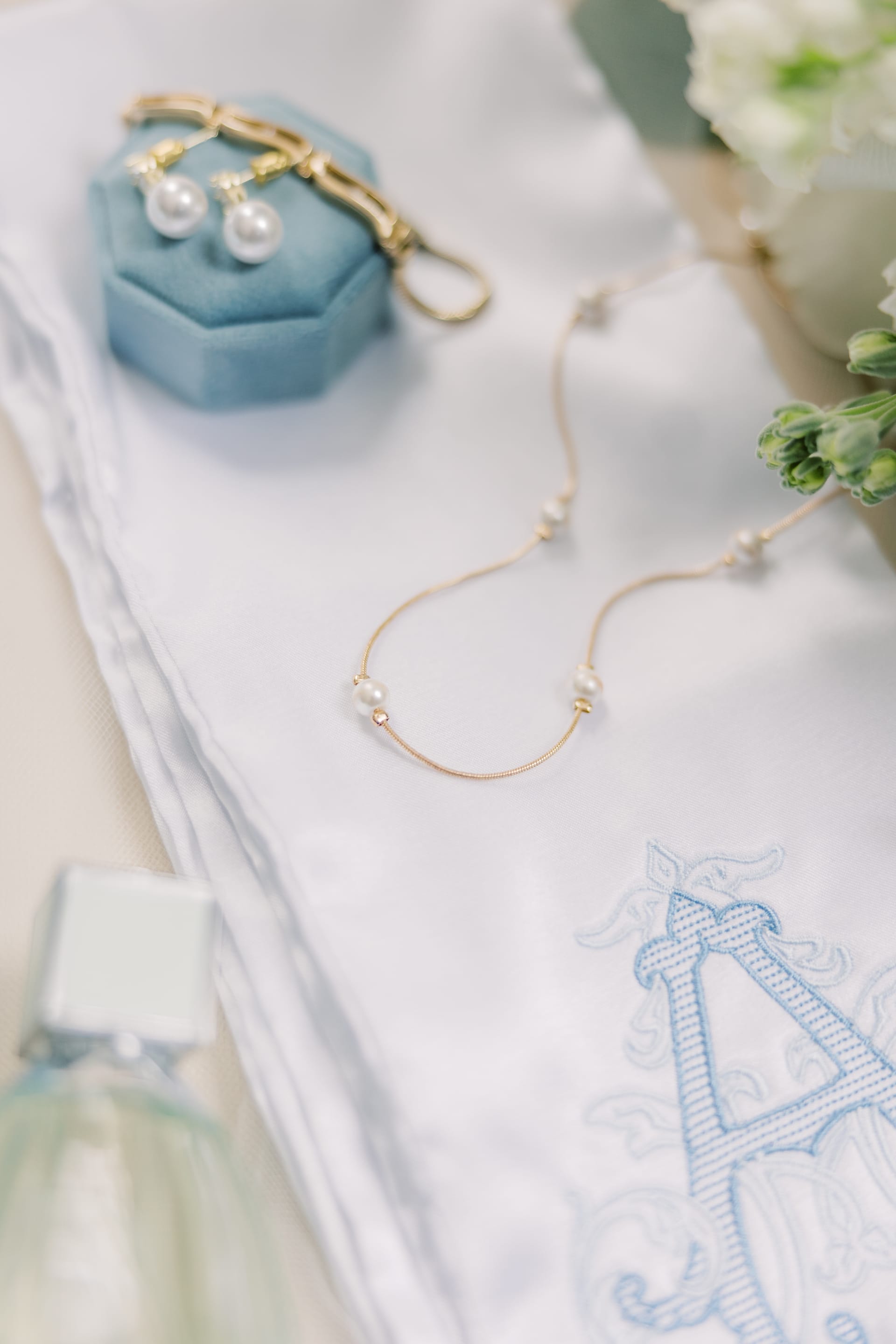 Gold pearl necklace and drop earrings in a blue velvet ring box arranged on embroidered white linen with flowers and perfume.