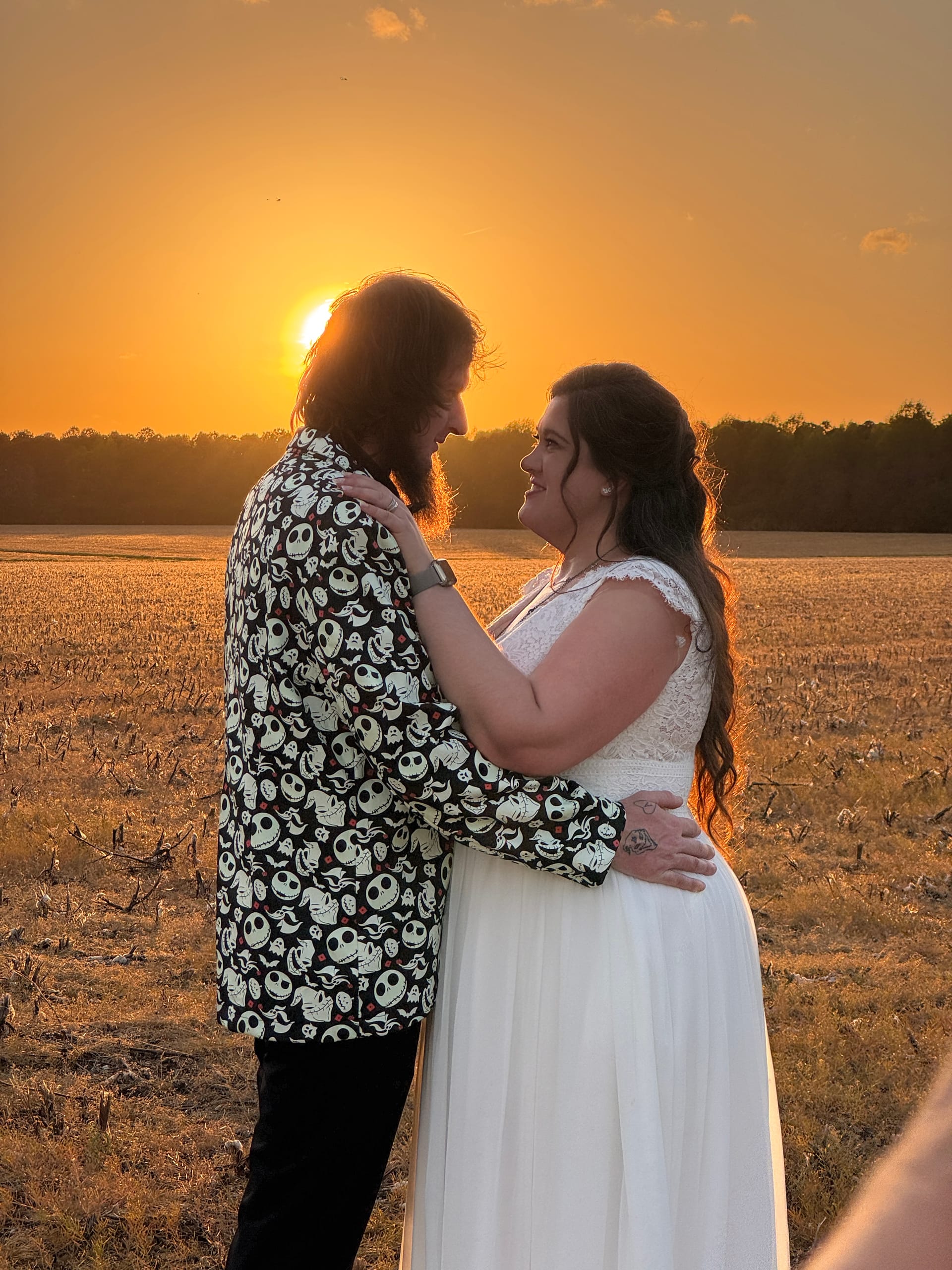 Bride in a white lace gown and groom in a patterned blazer share a romantic gaze at sunset over the golden farmland fields at Scarborough House.