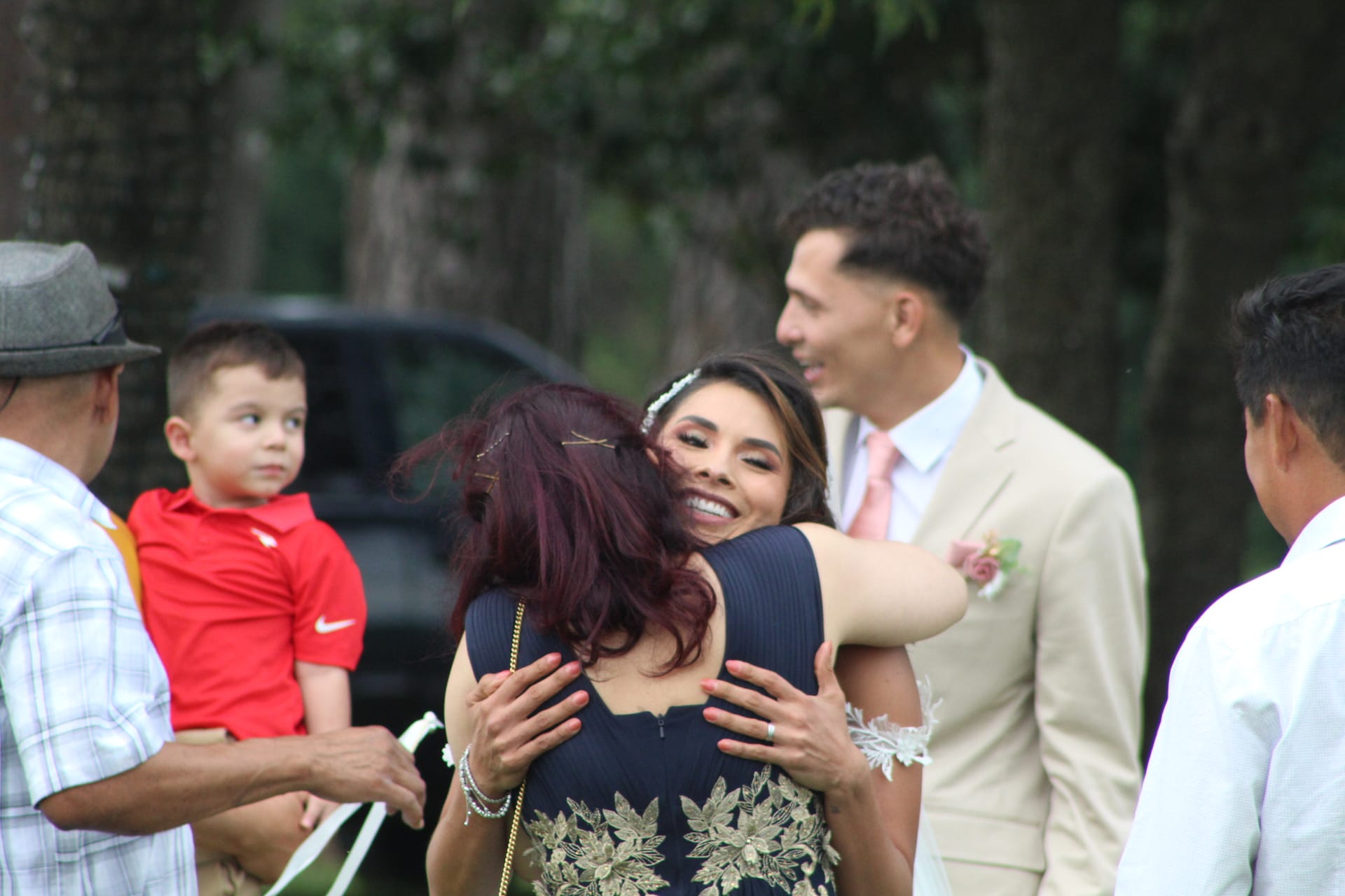 Smiling bride in a floral lace gown embraces a guest warmly while the groom in a tan suit looks on joyfully beneath oak trees at Scarborough House.