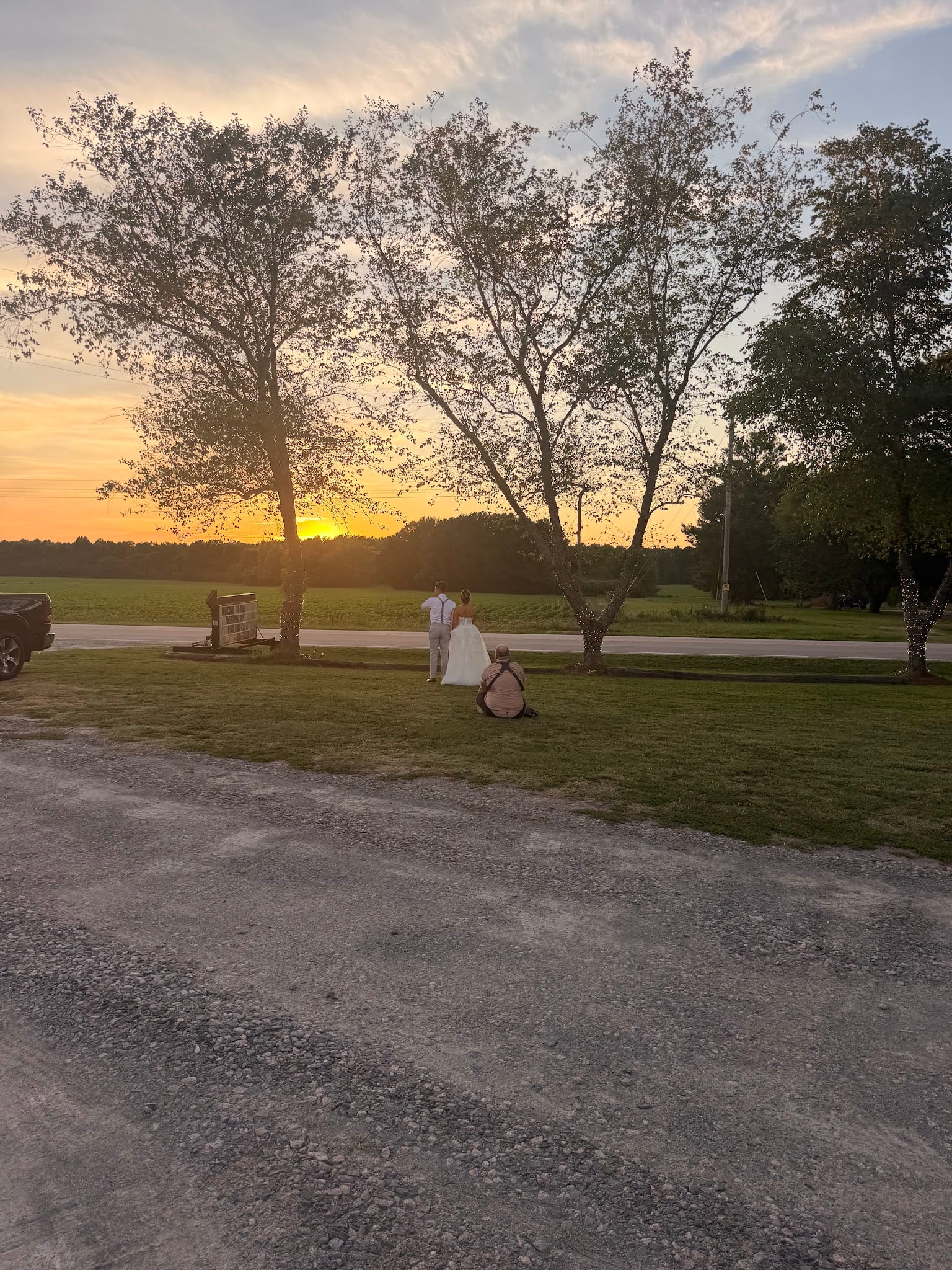 A bride in a white gown and groom stand together facing a glowing golden sunset over open farmland fields while a photographer captures their portrait at Scarborough House estate.