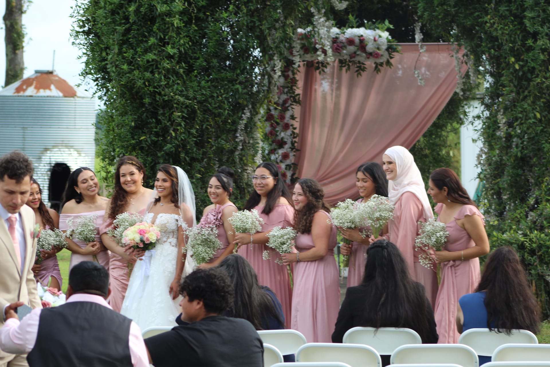 Smiling bride in white lace gown stands with her bridesmaids in blush pink dresses holding baby's breath bouquets beneath a floral-draped arch on the ceremony lawn at Scarborough House.
