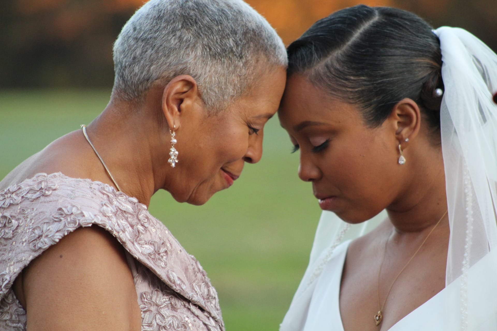A bride in a white gown and veil shares a tender forehead-to-forehead moment with an older woman in a blush lace dress against a soft green outdoor backdrop at Scarborough House.