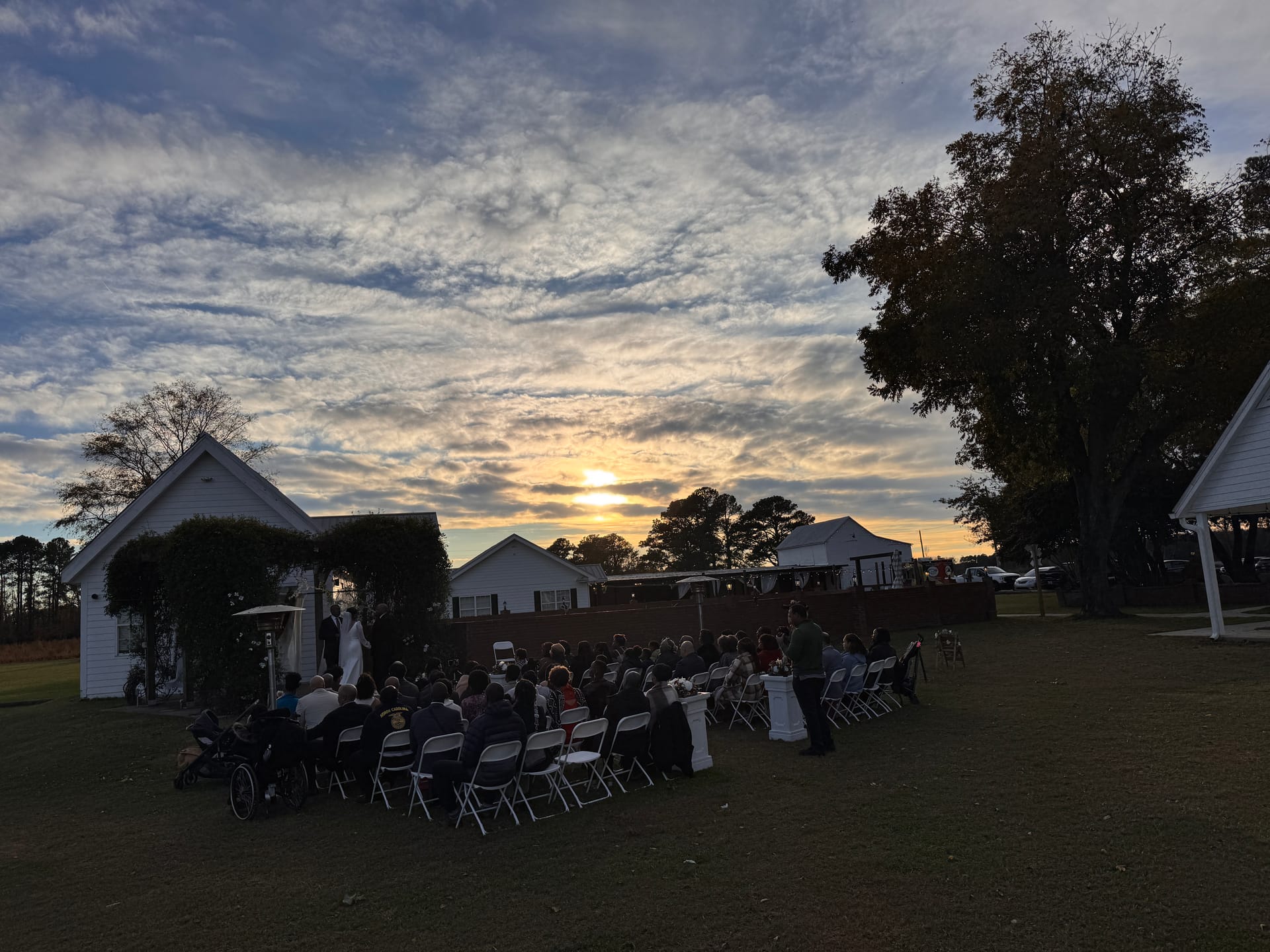 Outdoor wedding ceremony at dusk on the Scarborough House ceremony lawn with guests seated in white folding chairs as a golden sunset blazes through dramatic clouds on the horizon.