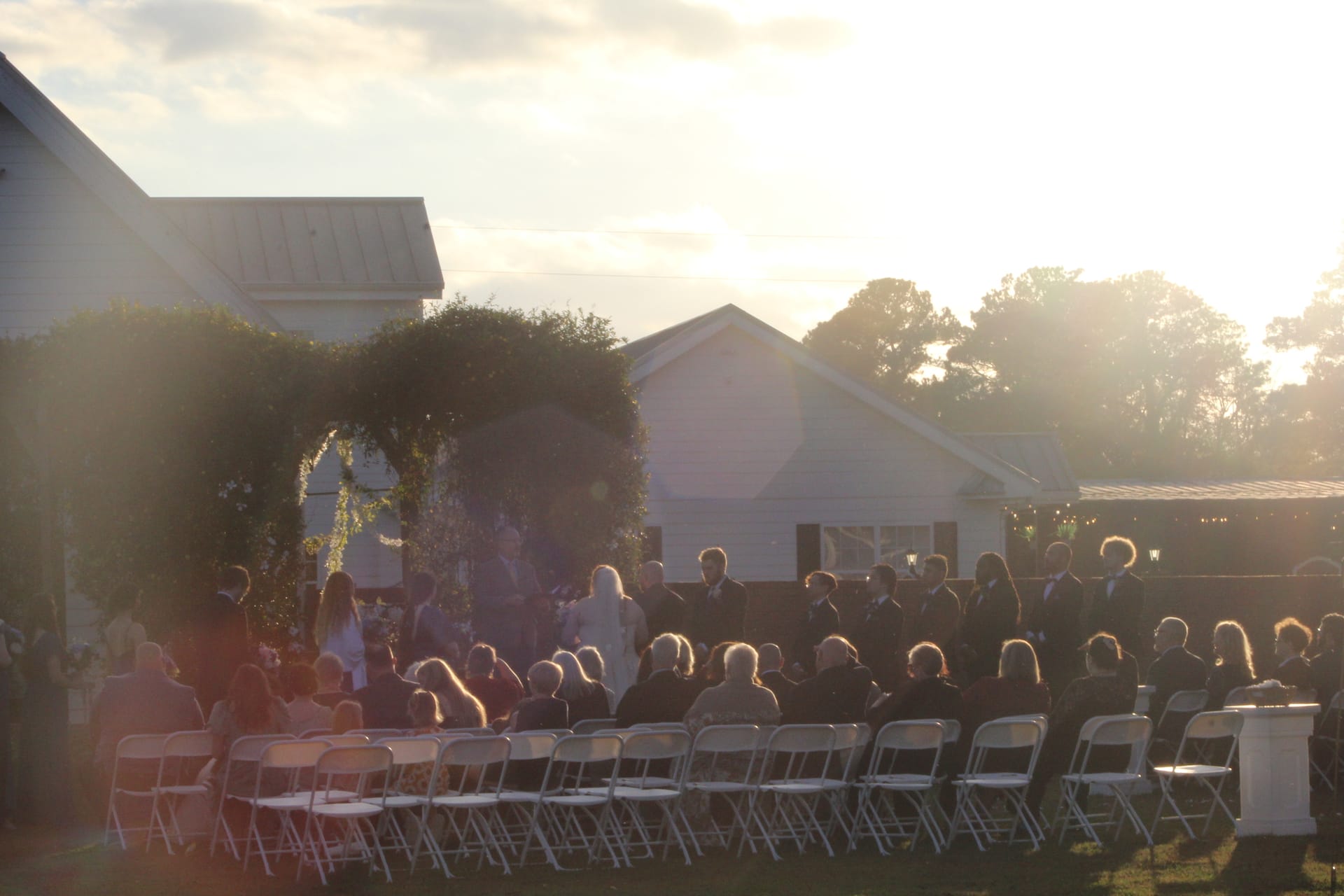 Guests seated in white folding chairs watch a wedding ceremony beneath a lush floral arch at golden hour on the Scarborough House ceremony lawn, with the historic white manor house glowing in the warm sunset light.