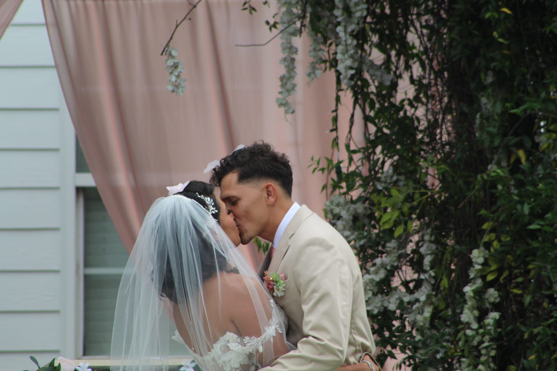 Bride in a lace gown and veil shares her first kiss with the groom in a tan suit beneath a blush-draped floral arch at Scarborough House.