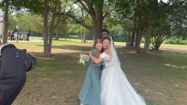 Bride in a white ball gown joyfully embracing a guest in a green floral dress beneath large oak trees at Scarborough House while a photographer captures the moment.