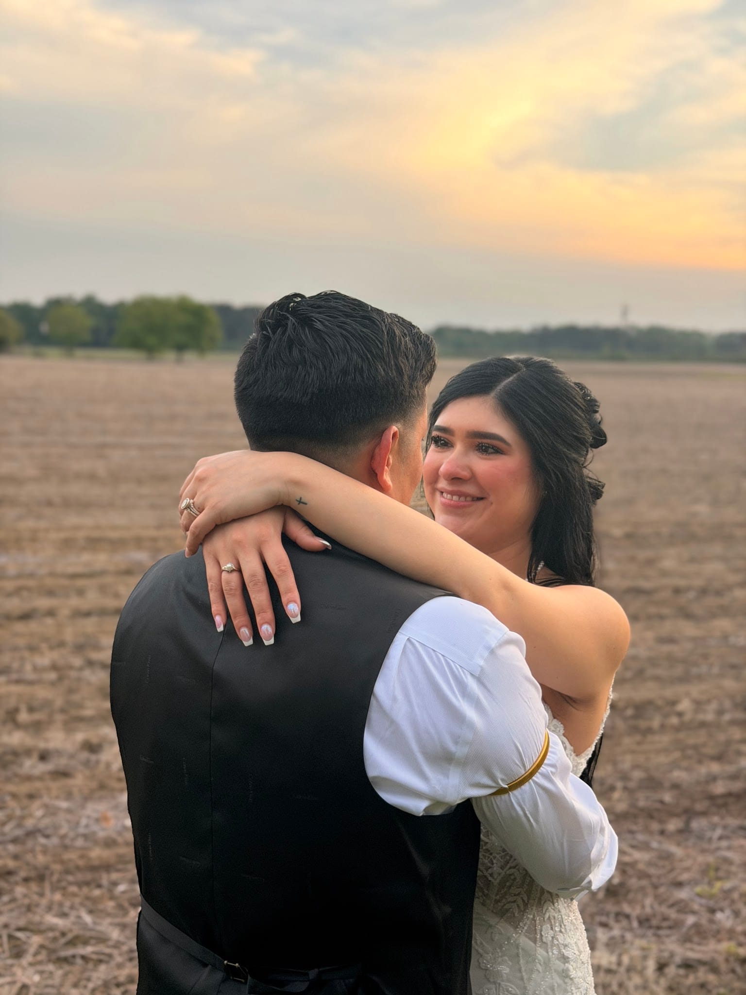 Bride smiles warmly while embracing her groom in an open farmland field at Scarborough House during a glowing golden-hour sunset.