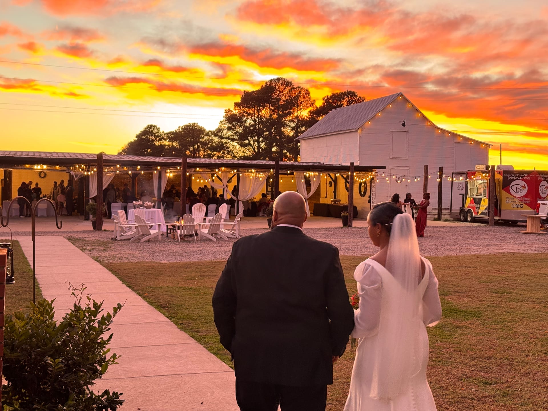 Bride in white gown and veil walks arm-in-arm with groom in black suit toward a warmly lit outdoor reception beneath a dramatic fiery sunset at Scarborough House wedding venue.