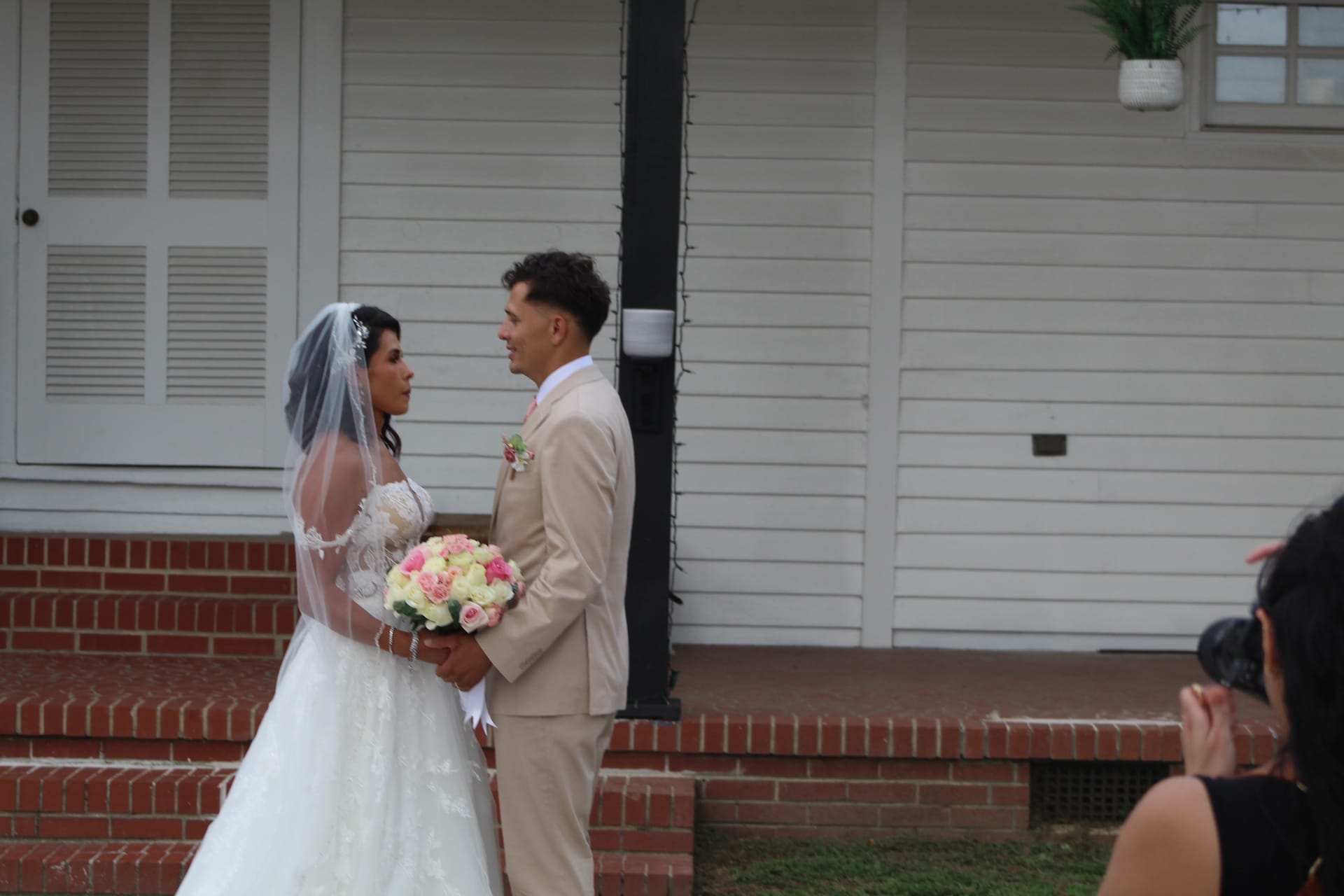 Bride in white lace gown holding a pink and cream rose bouquet shares a tender first-look moment with her groom in a tan suit outside the historic white clapboard facade of Scarborough House.