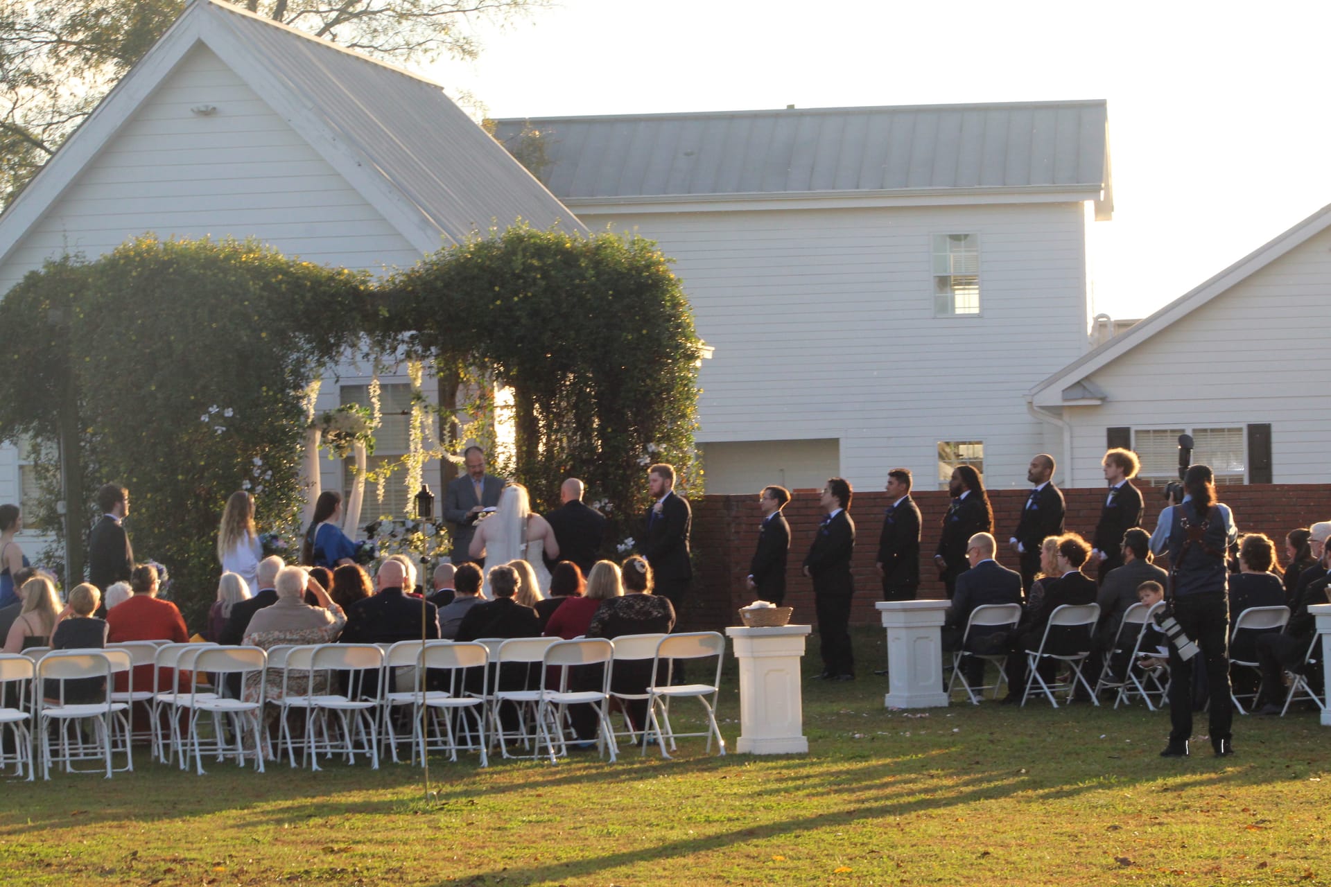 Outdoor wedding ceremony on the Scarborough House lawn at golden hour with guests seated in white chairs facing the couple under a lush floral arch, groomsmen standing to the right beside white pedestal arrangements.