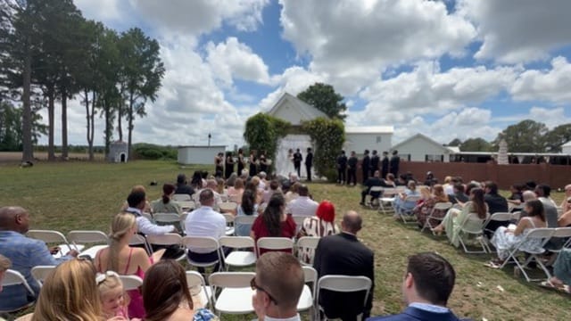 Guests seated in white folding chairs on the ceremony lawn watch a wedding party gathered at a floral arch in front of a white chapel at Scarborough House historic estate.