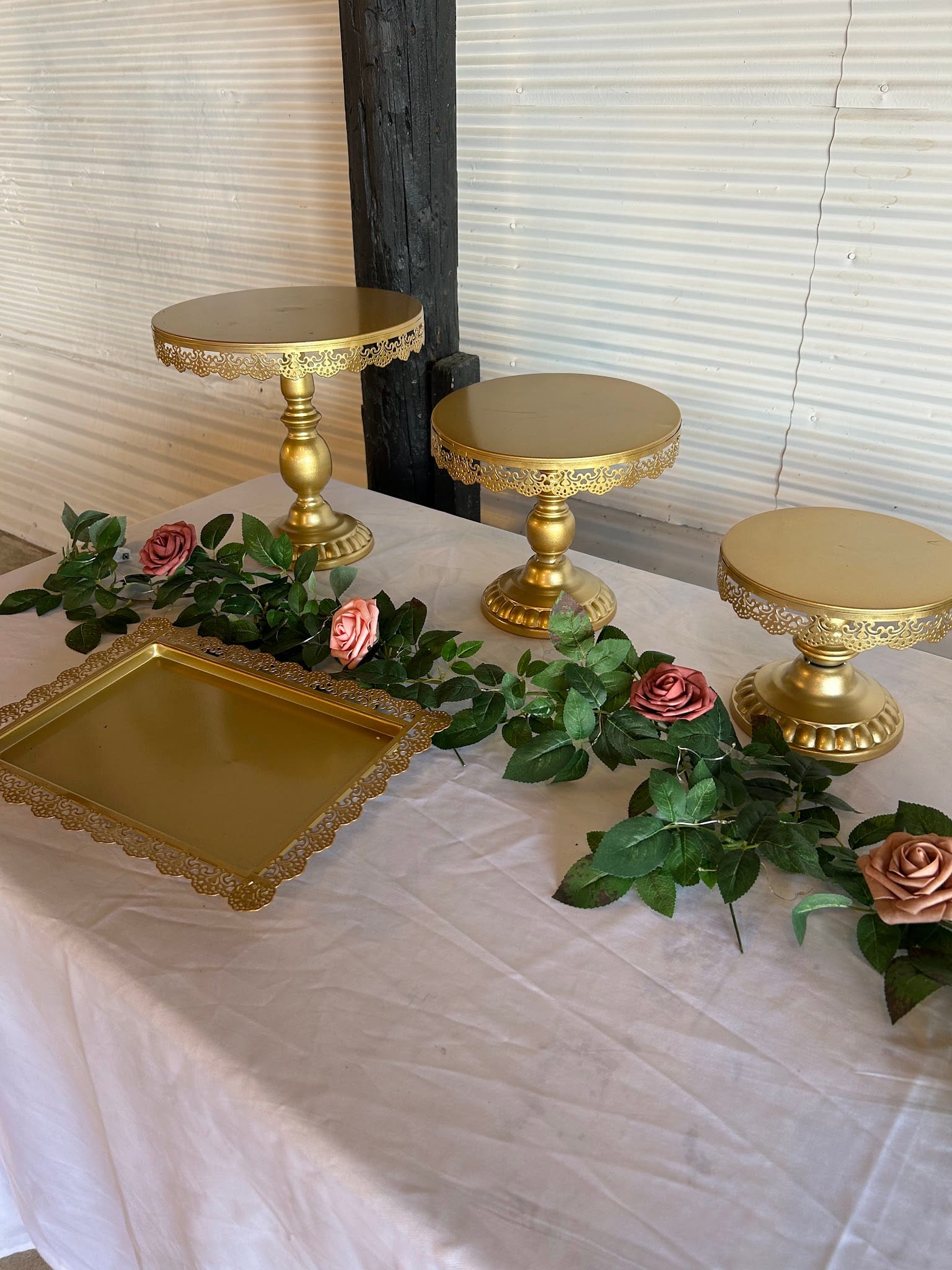 Three ornate gold pedestal cake stands with lace-edged detailing and a matching gold tray arranged on a white linen table with pink roses and green garland at Scarborough House.