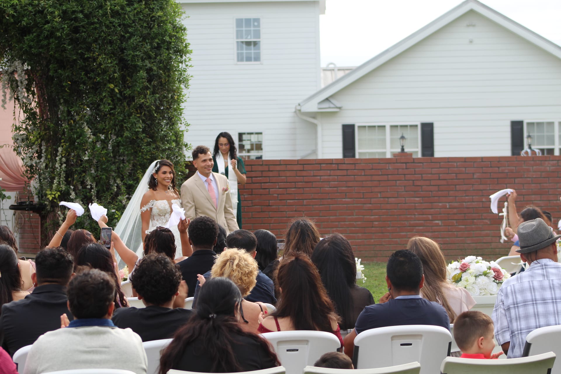 Bride in a lace gown and groom in a tan suit walk the outdoor ceremony aisle as seated guests wave white handkerchiefs in celebration at Scarborough House estate.