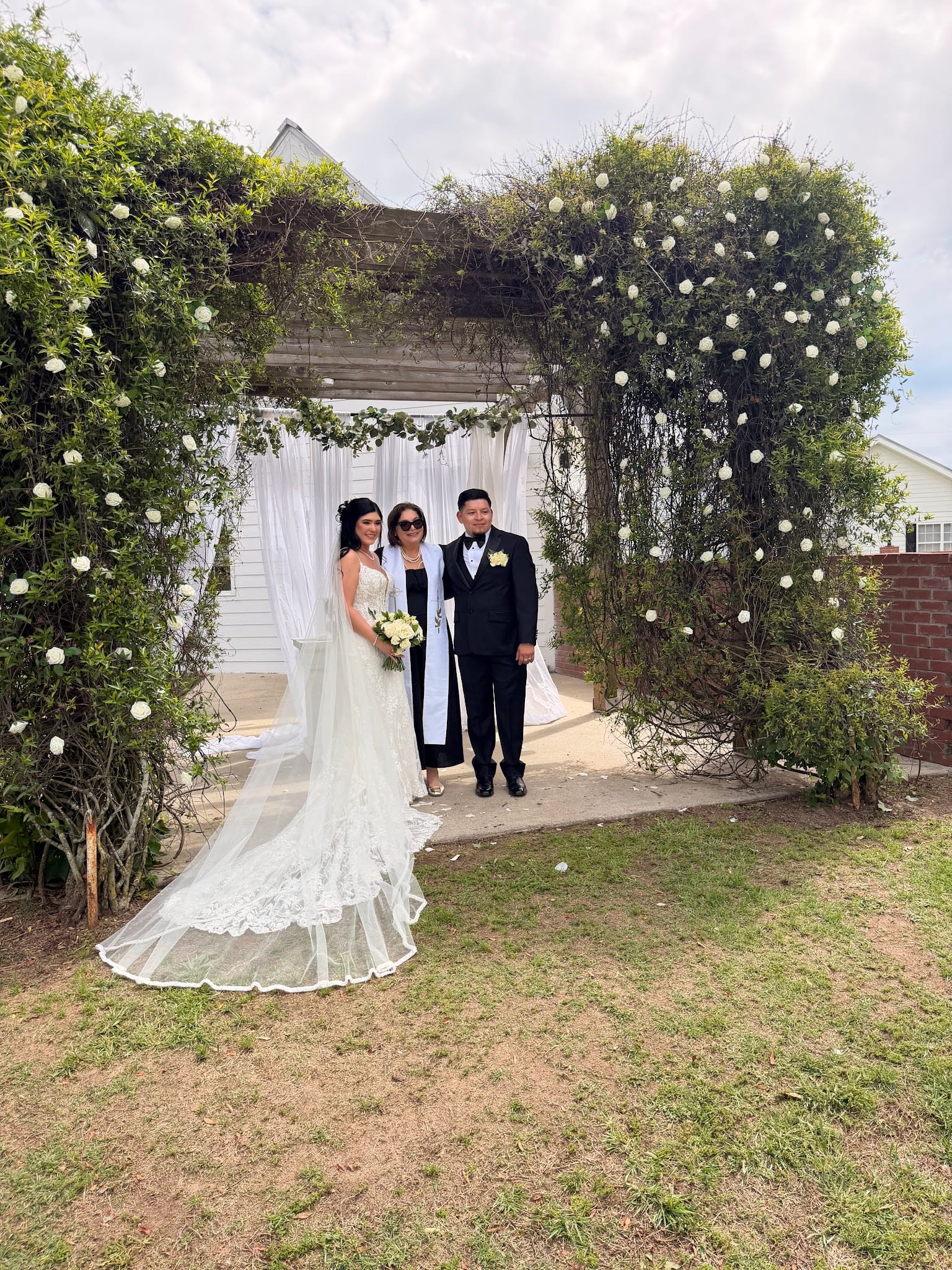 Bride in a lace gown with long veil and groom in a black tuxedo pose with their officiant beneath a vine-covered archway adorned with white roses at Scarborough House historic estate.