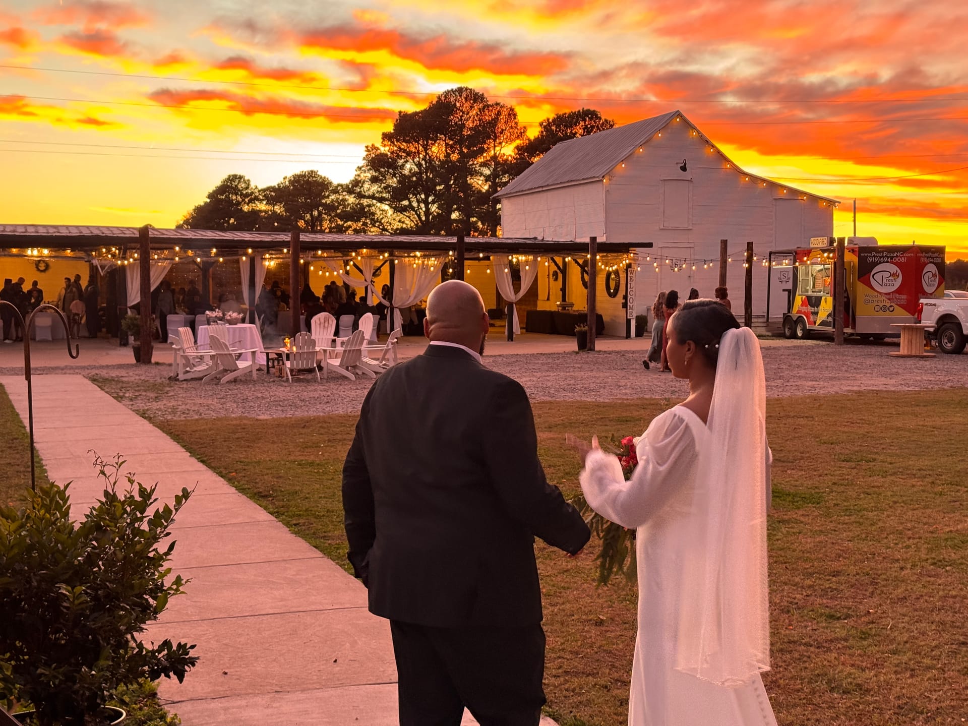 Bride in a white gown and veil walks with her partner in a dark suit toward a warmly lit outdoor barn wedding reception beneath a vivid sunset sky.