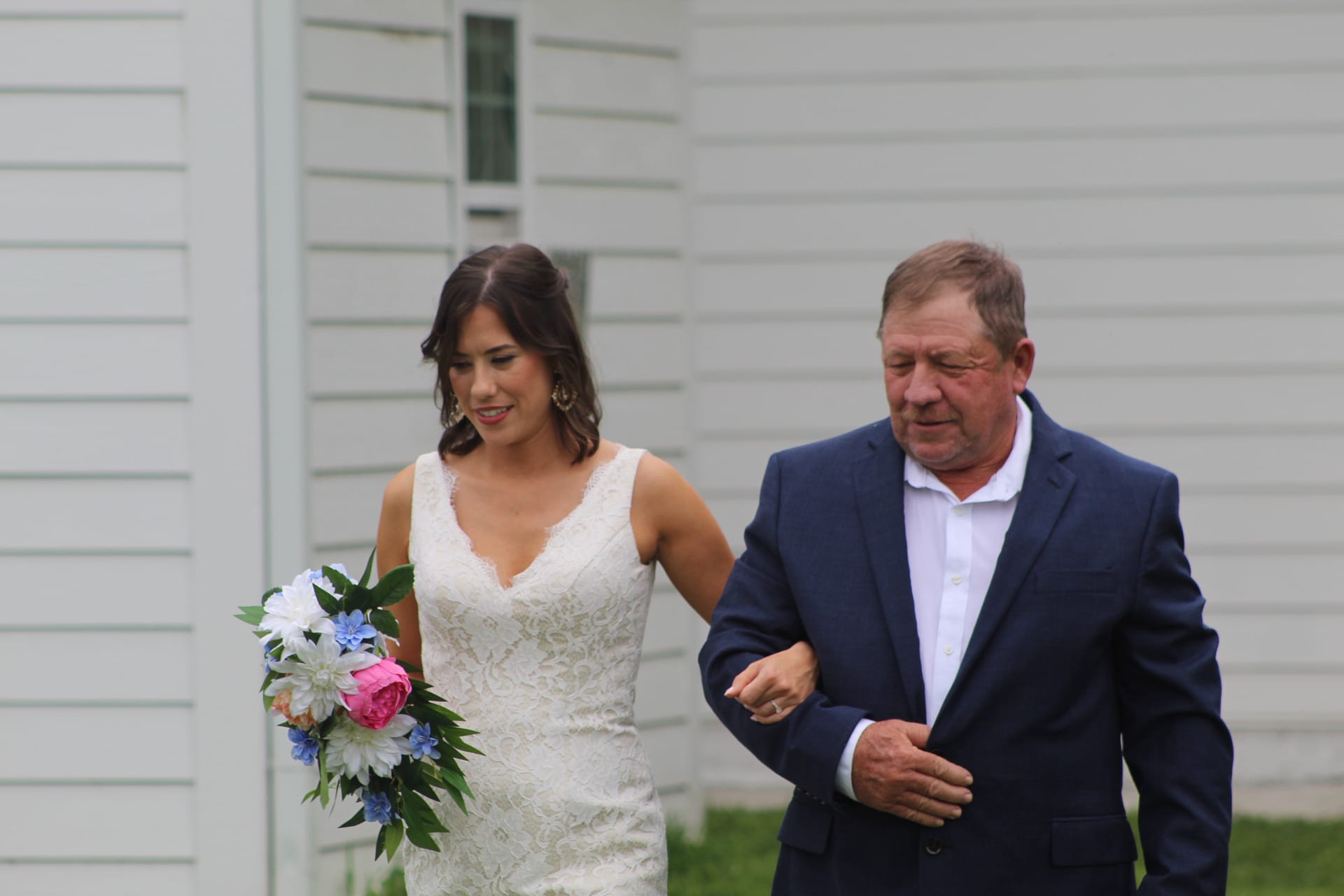 A smiling bride in a white lace gown holding a pink and blue bouquet walks arm-in-arm with an older gentleman in a navy suit outside Scarborough House during the processional.
