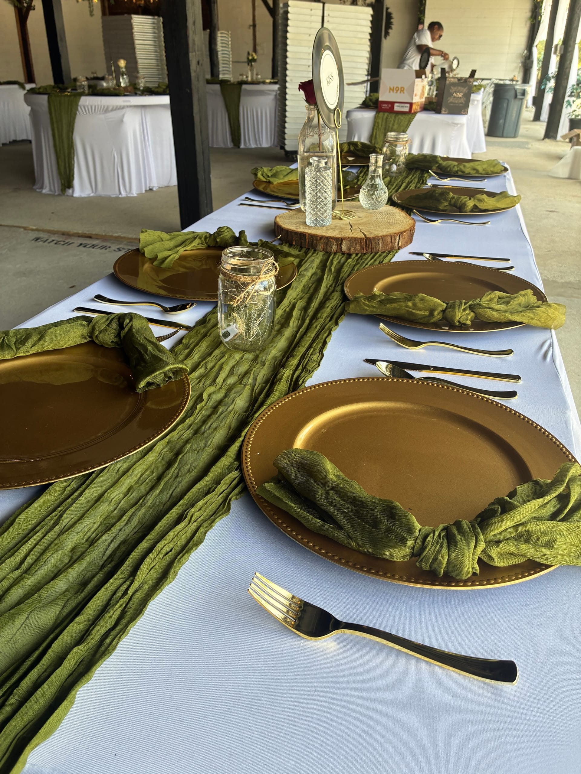 Elegantly set reception table at Scarborough House featuring gold charger plates, knotted olive green gauze napkins, gold flatware, mason jar centerpieces, and a rustic wood-slice display on a white linen tablecloth.
