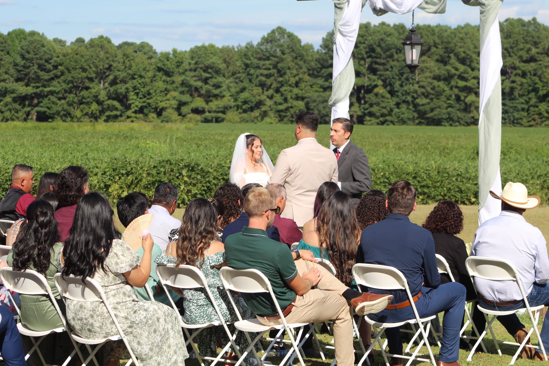Wedding guests seated in white folding chairs watch as a bride in a white veil and groom exchange vows beneath a draped arch on a sunlit ceremony lawn surrounded by green farmland fields.