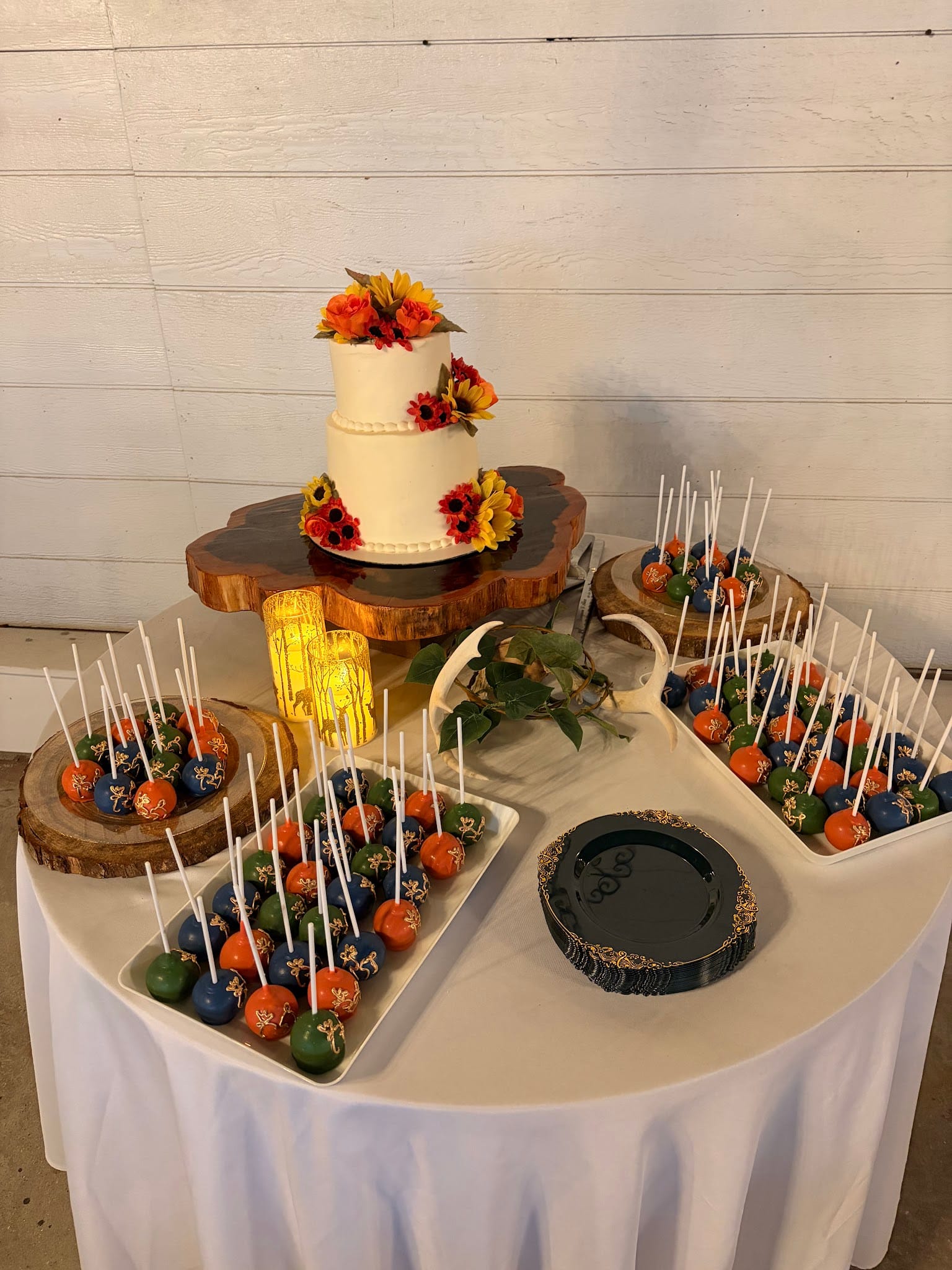 Elegant wedding dessert table featuring a two-tier white cake adorned with autumn sunflowers and a colorful array of orange, blue, and green cake pops displayed on rustic wood slab risers.