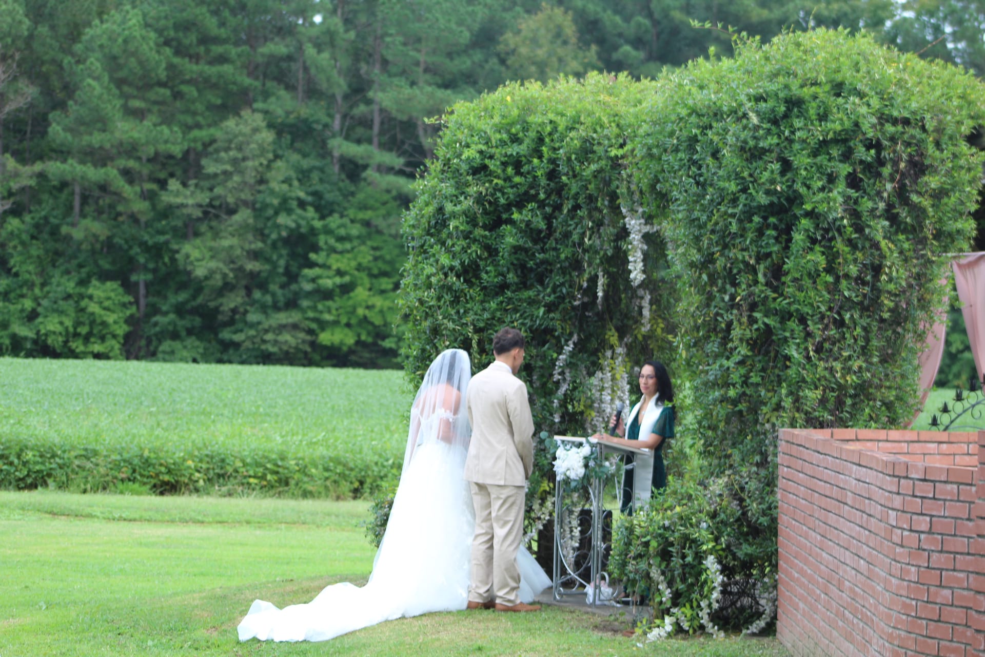 Bride and groom stand before an officiant at a lush ivy-covered archway during an outdoor wedding ceremony at Scarborough House, with open farmland fields and tall trees in the background.