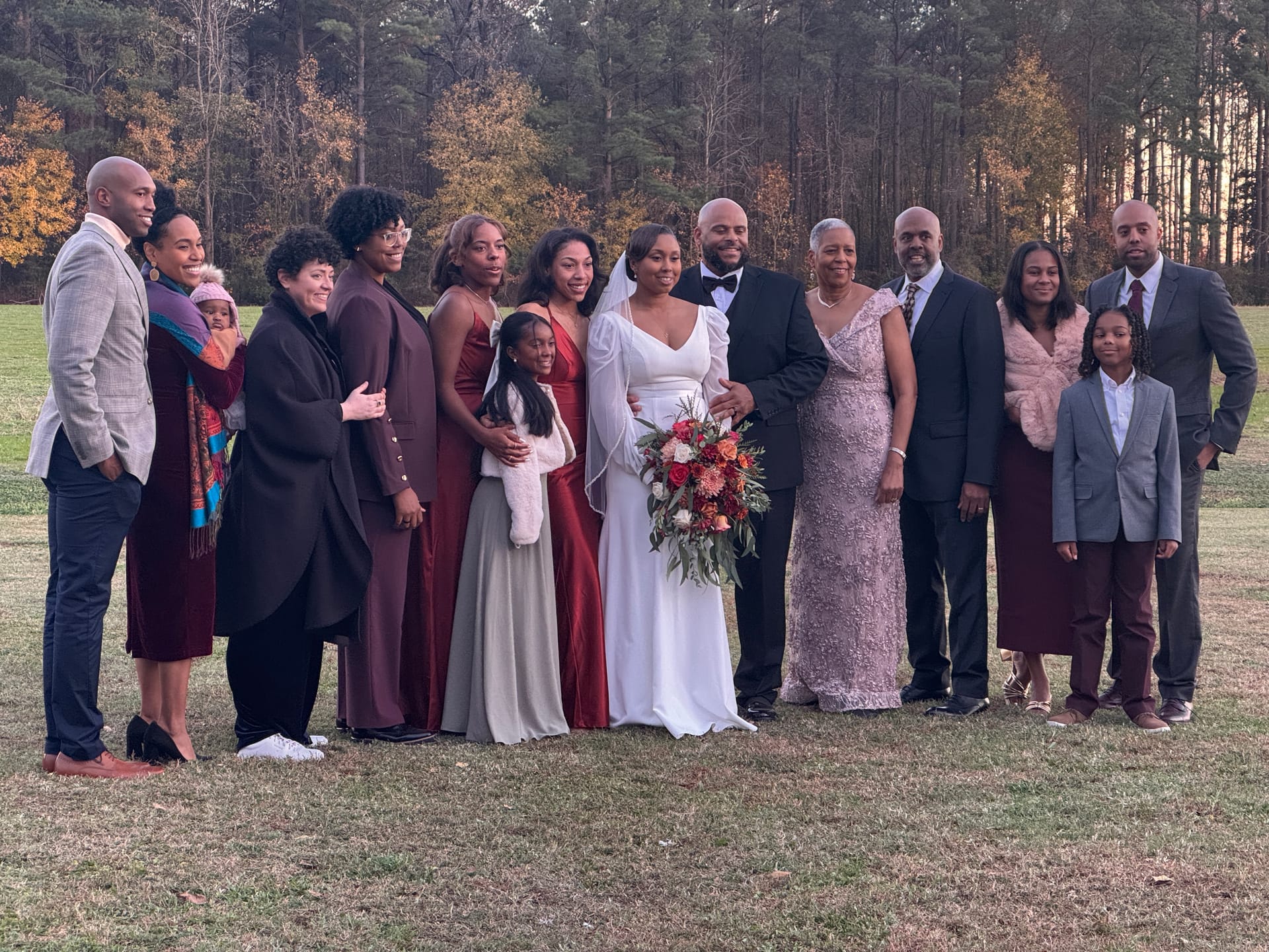Bride in a white gown with veil and groom in a black tuxedo surrounded by a large extended family group on the grassy ceremony lawn at Scarborough House, with pine trees glowing in autumn light behind them.